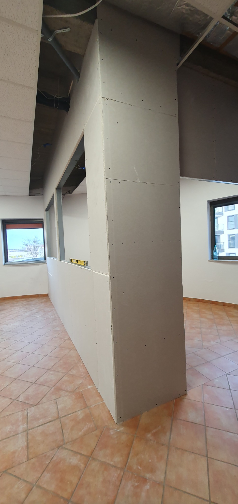 Montaż ścianki działowej z płyt karton-gips w biurze. Widoczne profile, płyty KG, poziomica. Pomieszczenie z oknami i terakotą. Sufit podwieszany w trakcie montażu.