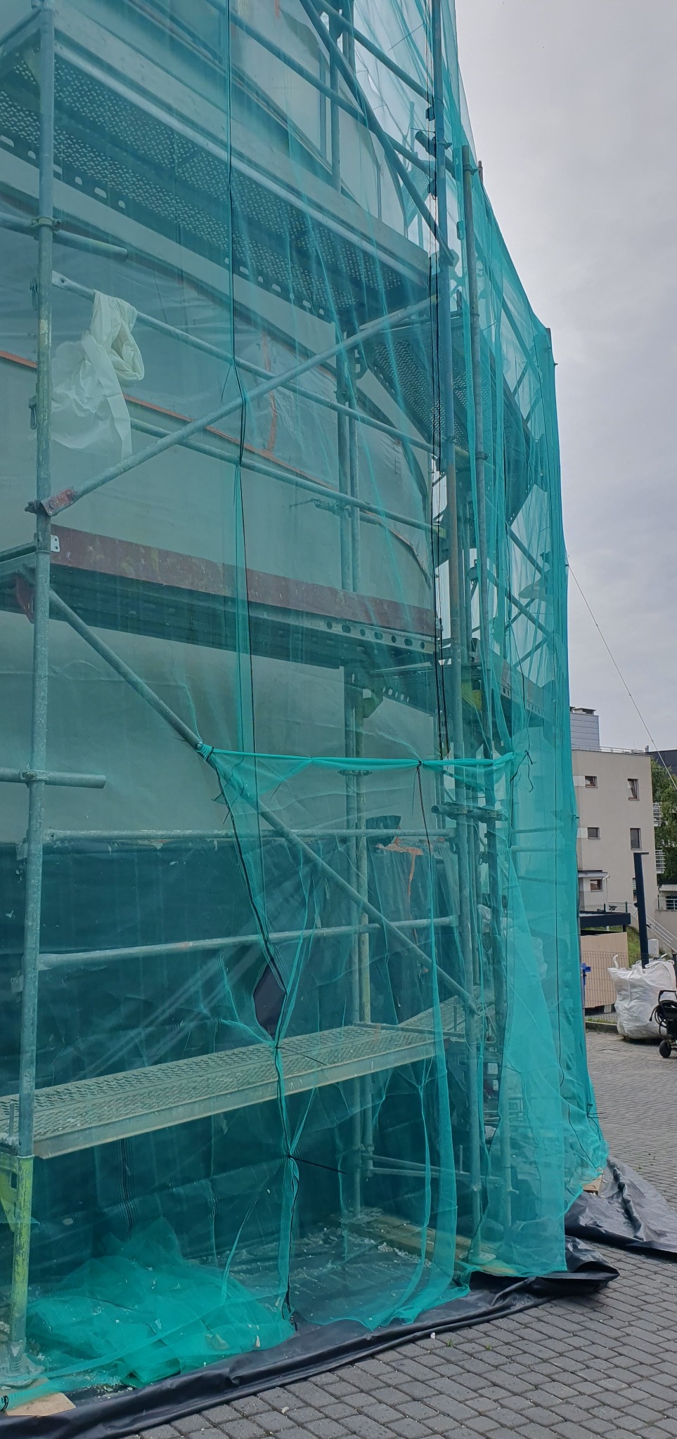 Rusztowanie z zieloną siatką ochronną na tle budynku w trakcie remontu. Widoczne elementy konstrukcyjne i fragmenty elewacji. Ujęcie z dołu, podkreślające wysokość rusztowania.