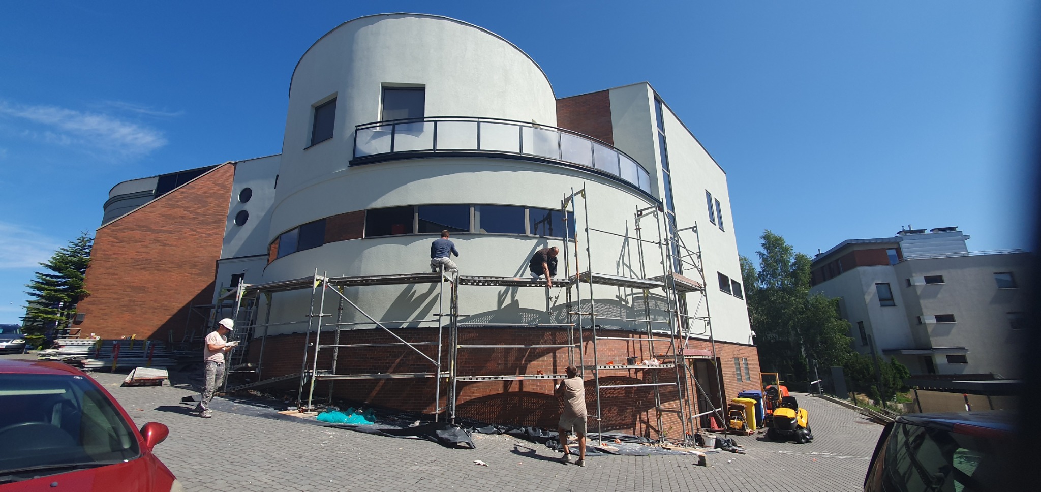 Praca na rusztowaniu przy budynku o nietypowej architekturze. Dwóch pracowników maluje elewację. Widoczny fragment czerwonego samochodu i traktor.