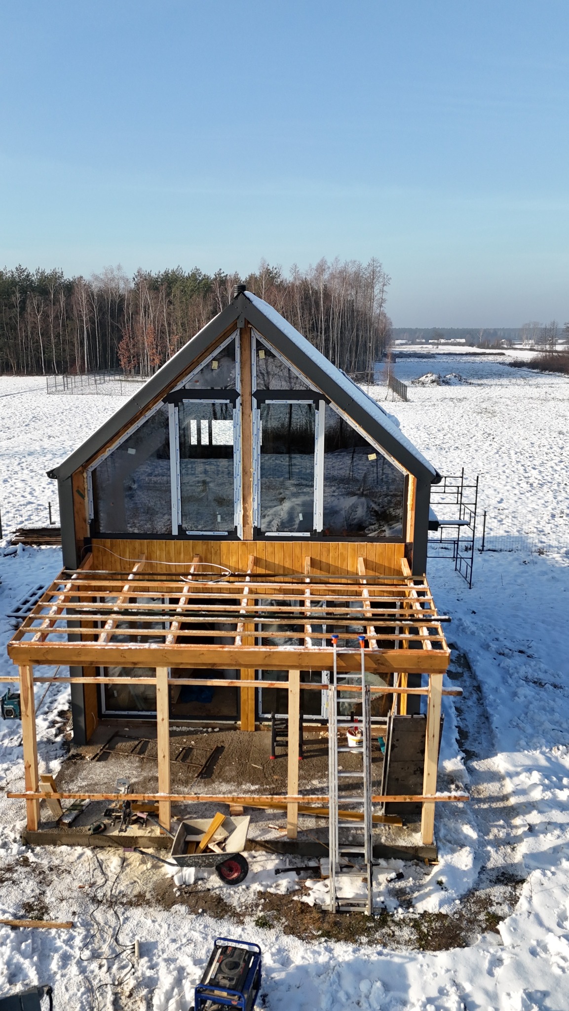 Budowa domu z dużymi oknami w zimowym krajobrazie. Widoczne rusztowania i narzędzia, sugerujące etap prac wykończeniowych. Jasne, naturalne światło.