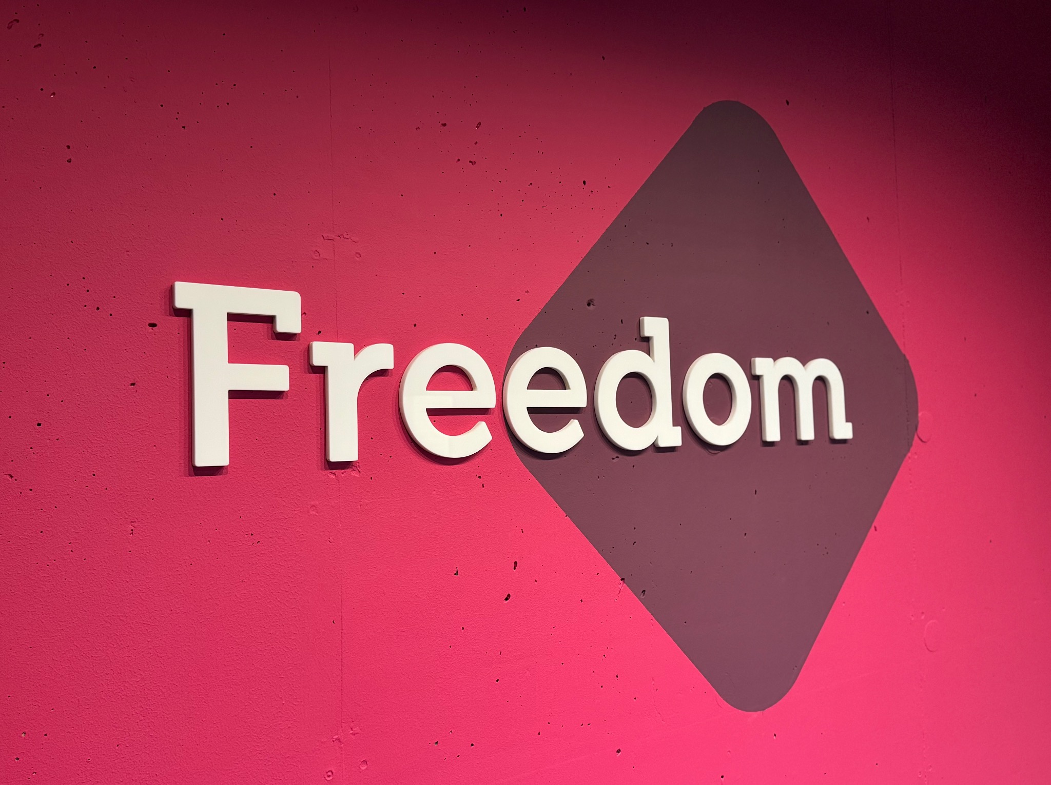 Biały napis 'Freedom' na różowo-fioletowym tle z geometrycznym kształtem. Minimalistyczny design, nowoczesny branding, wyraźny kontrast kolorów, bliskie ujęcie.