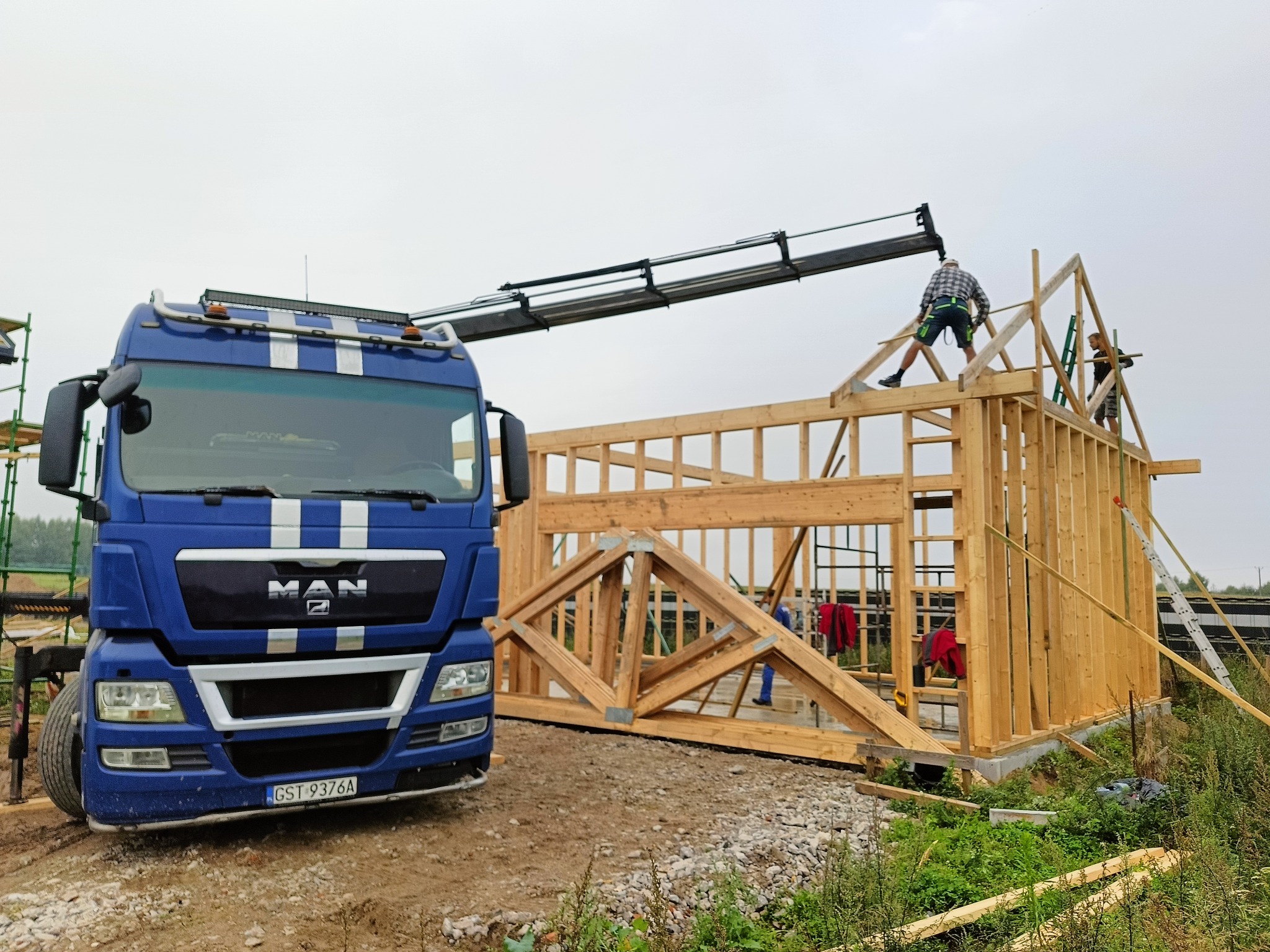 Niebieska ciężarówka MAN z dźwigiem transportuje elementy drewniane konstrukcji szkieletowej domu. Dwóch pracowników na dachu. Etap budowy.