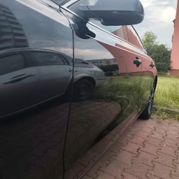 Dawid Salicki - Błyszczący, czarny samochód po polerowaniu, z wyraźnym odbiciem otoczenia w lakierze. Widoczny fragment karoserii, lusterko i felga. Tło: budynek i zieleń.