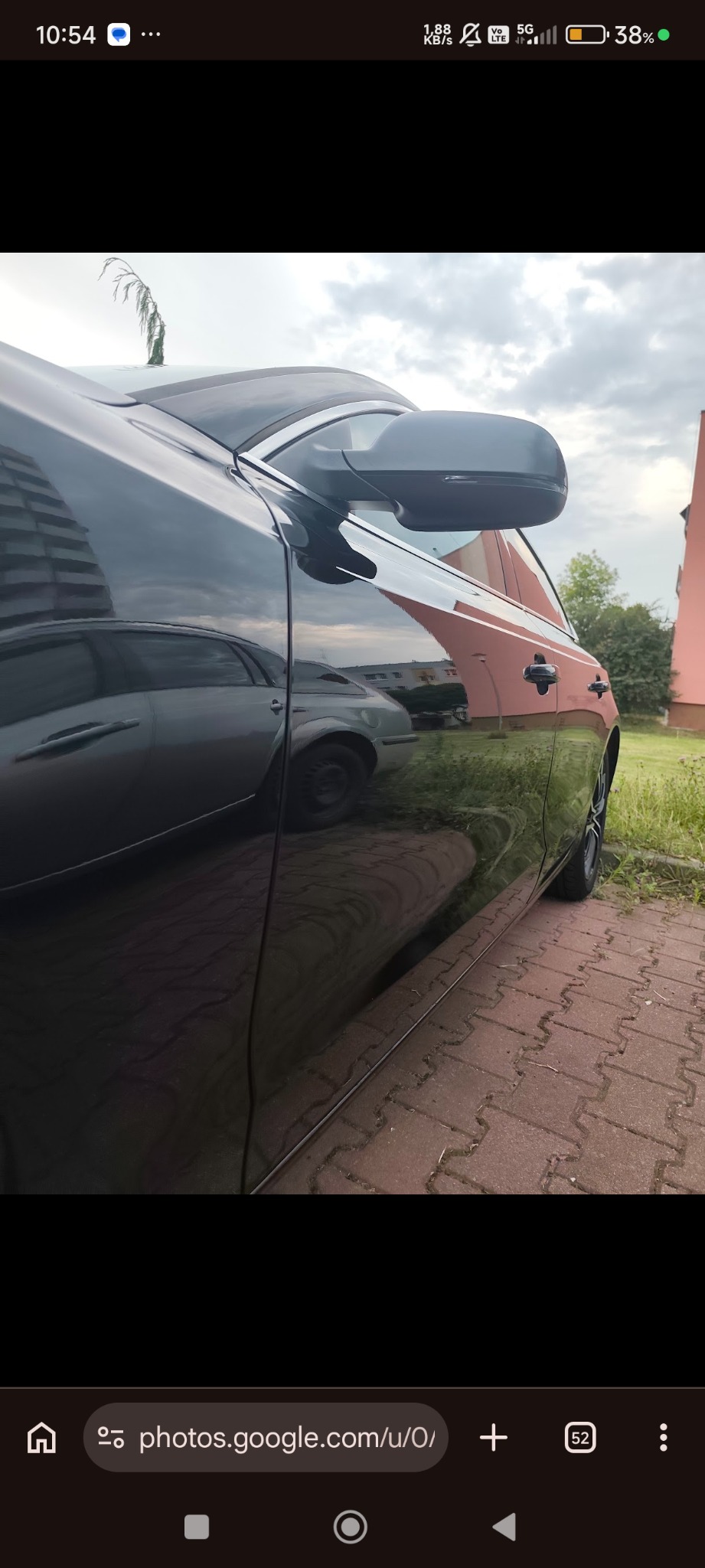 Błyszczący, czarny samochód po polerowaniu, z wyraźnym odbiciem otoczenia w lakierze. Widoczny fragment karoserii, lusterko i felga. Tło: budynek i zieleń.