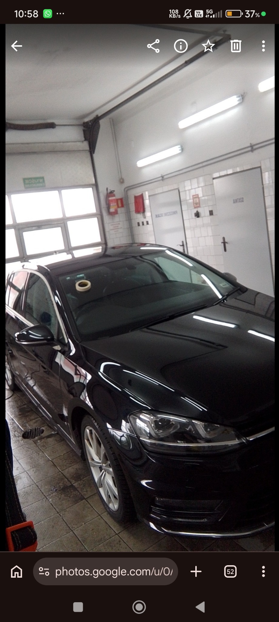 Błyszczący, czarny samochód w warsztacie blacharsko-lakierniczym, z taśmą maskującą na dachu. Widoczne płytki na ścianach i podłodze, oświetlenie warsztatowe.