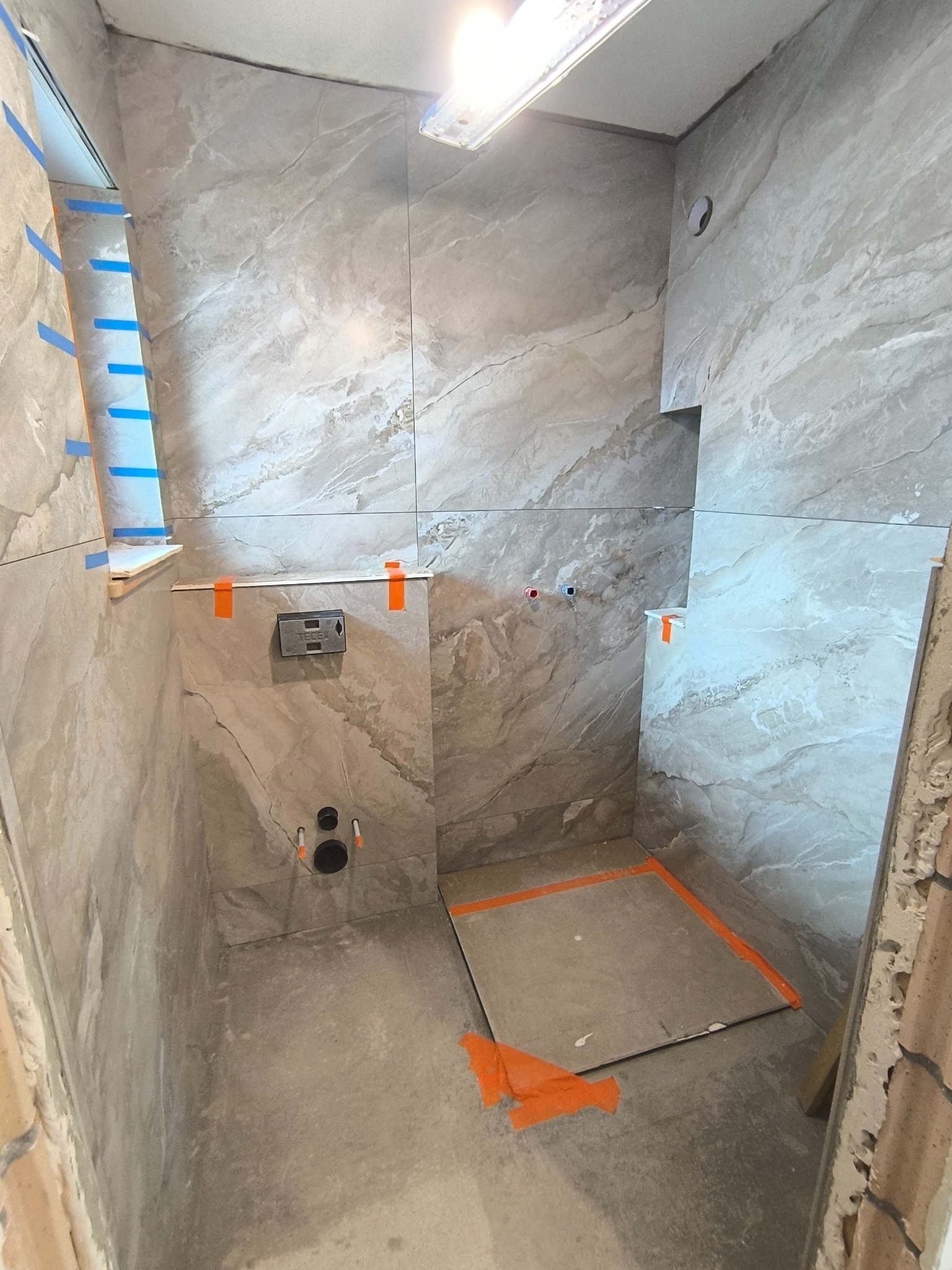 Wnętrze łazienki w trakcie prac wykończeniowych. Widoczne szare płytki imitujące marmur na ścianach, przygotowane miejsce na brodzik i instalacje sanitarne.