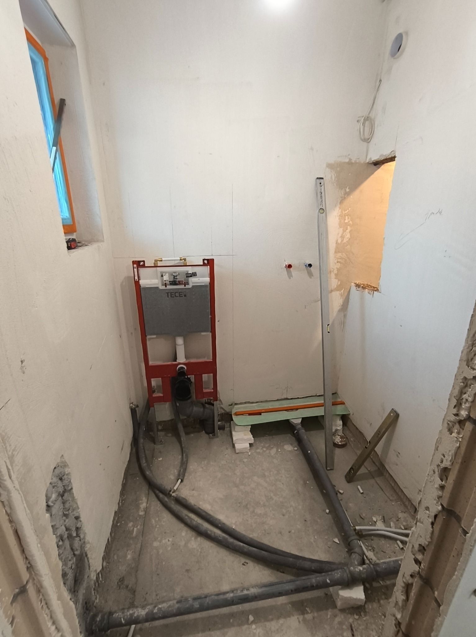 Montaż stelaża podtynkowego TECE w surowej łazience. Widoczne rury, poziomica i okno z niebieską folią. Ściany w trakcie przygotowania do wykończenia.