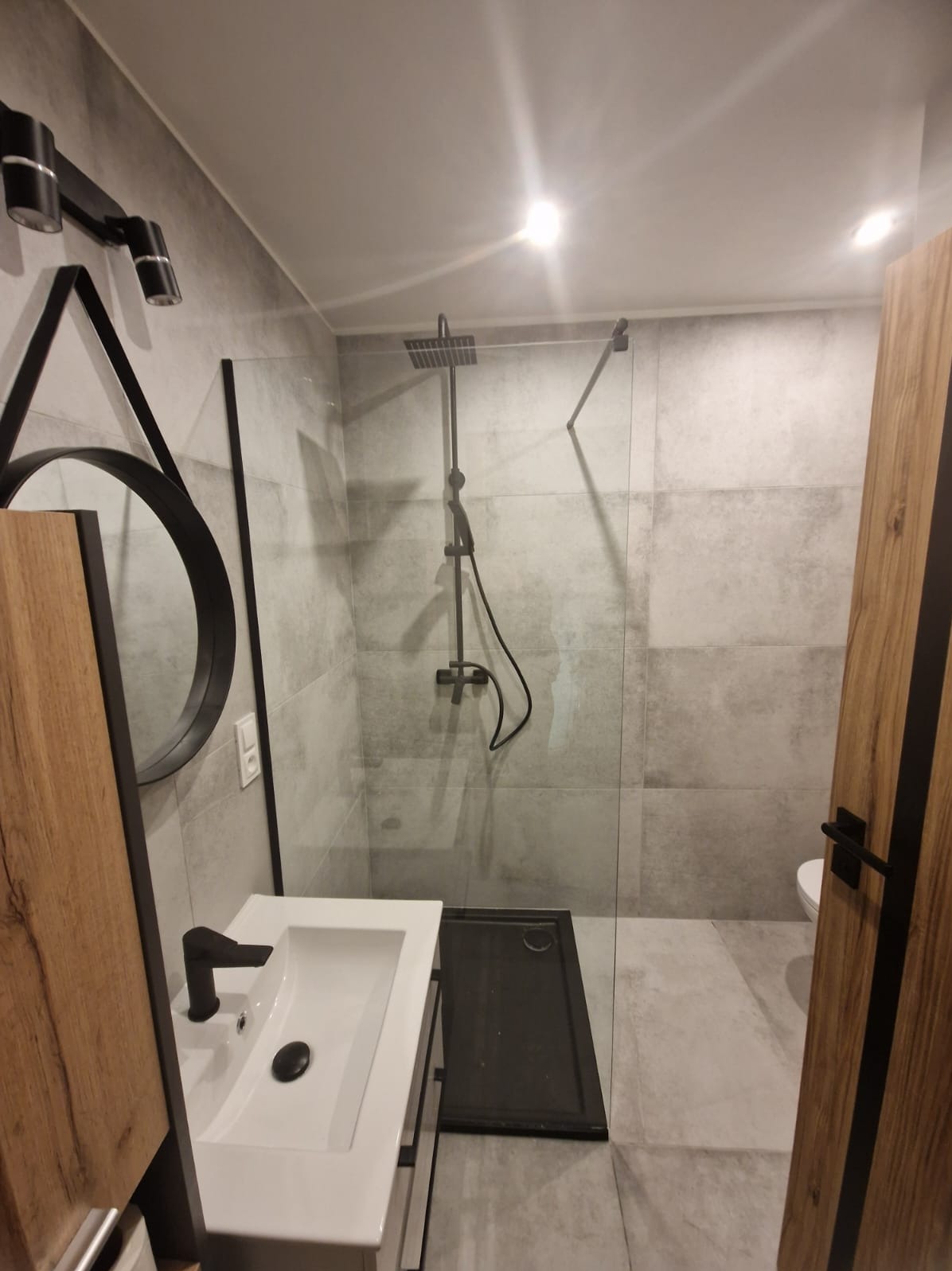 Nowoczesna łazienka z kabiną prysznicową typu walk-in, umywalką z czarną armaturą i okrągłym lustrem z geometrycznym akcentem. Szare płytki i drewniane wykończenia.
