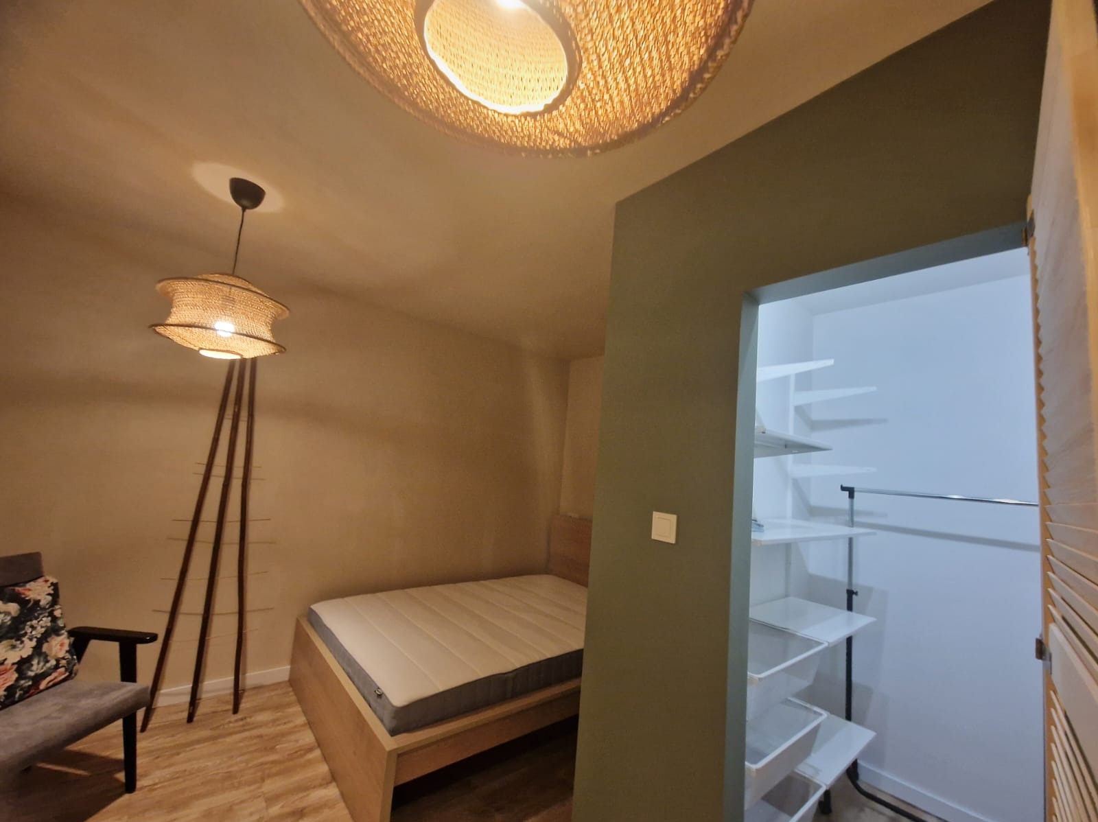 Jasne wnętrze sypialni z łóżkiem, lampą z abażurem i otwartą garderobą z półkami. Ściany w odcieniach beżu i oliwki. Podłoga z paneli imitujących drewno.