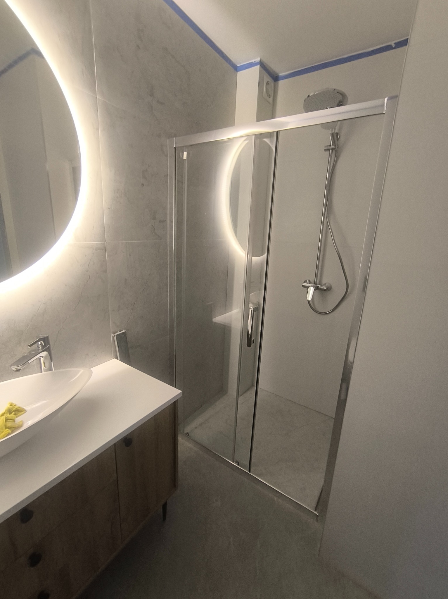 Wykończona łazienka z kabiną prysznicową, umywalką na szafce z drewnianymi frontami i okrągłym lustrem z podświetleniem LED. Szare płytki na ścianach i podłodze.