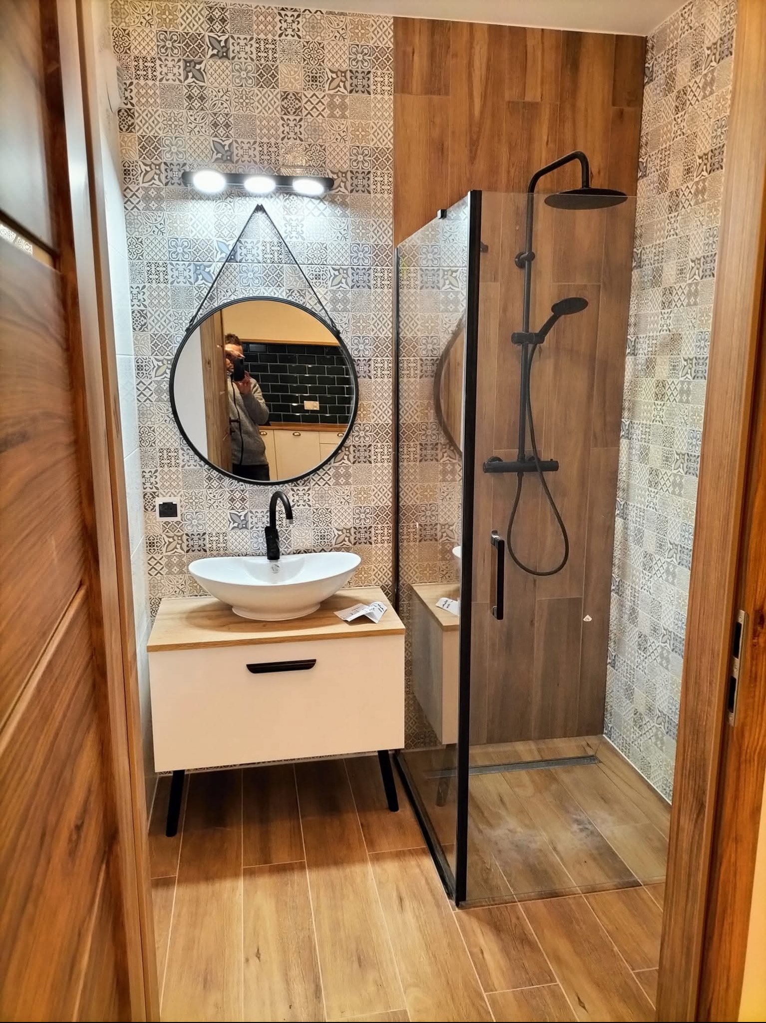 Nowoczesna łazienka z kabiną prysznicową typu walk-in, umywalką nablatową na szafce i okrągłym lustrem z oświetleniem. Płytki w geometryczne wzory na ścianach.