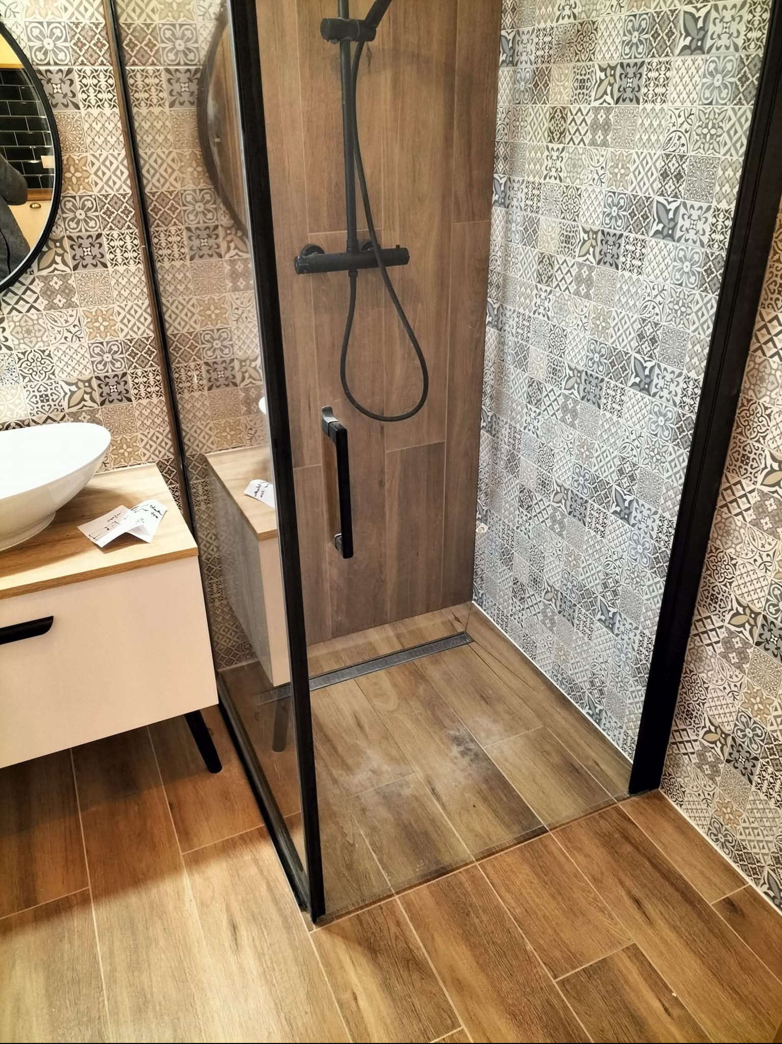 Nowoczesna kabina prysznicowa z czarnymi ramami i deszczownicą, drewnopodobne płytki podłogowe i wzorzyste płytki ścienne. Umywalka nablatowa z szafką.