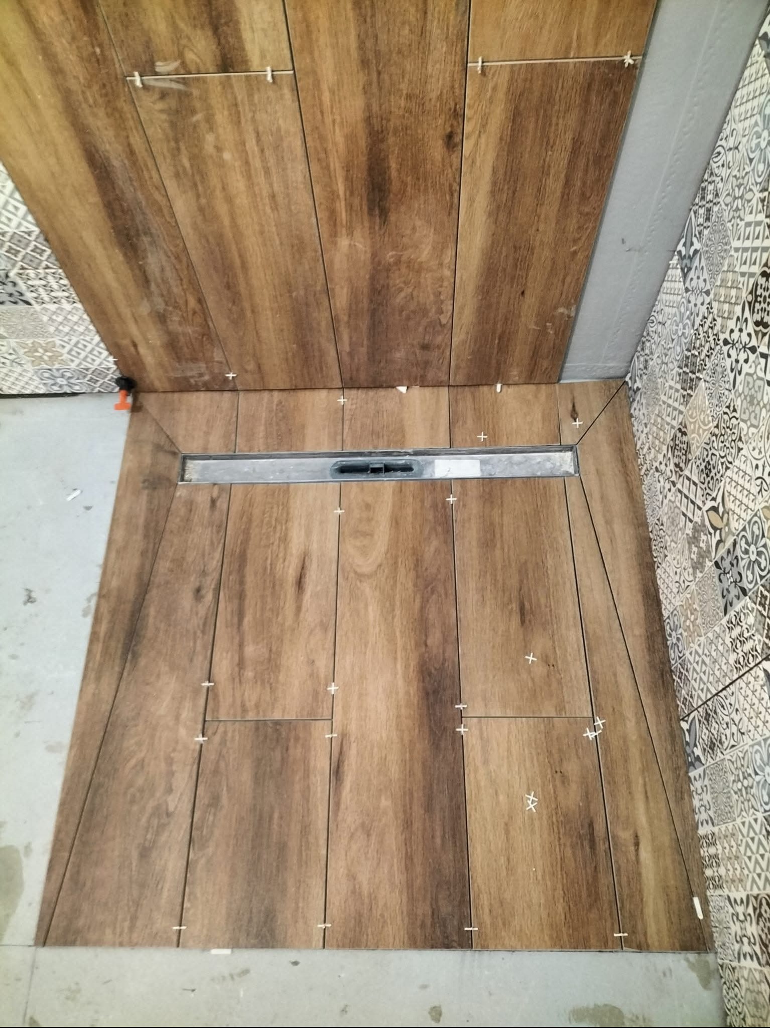 Układanie płytek imitujących drewno w kabinie prysznicowej z odpływem liniowym. Widoczne krzyżyki dystansowe i wzorzyste płytki na ścianie.