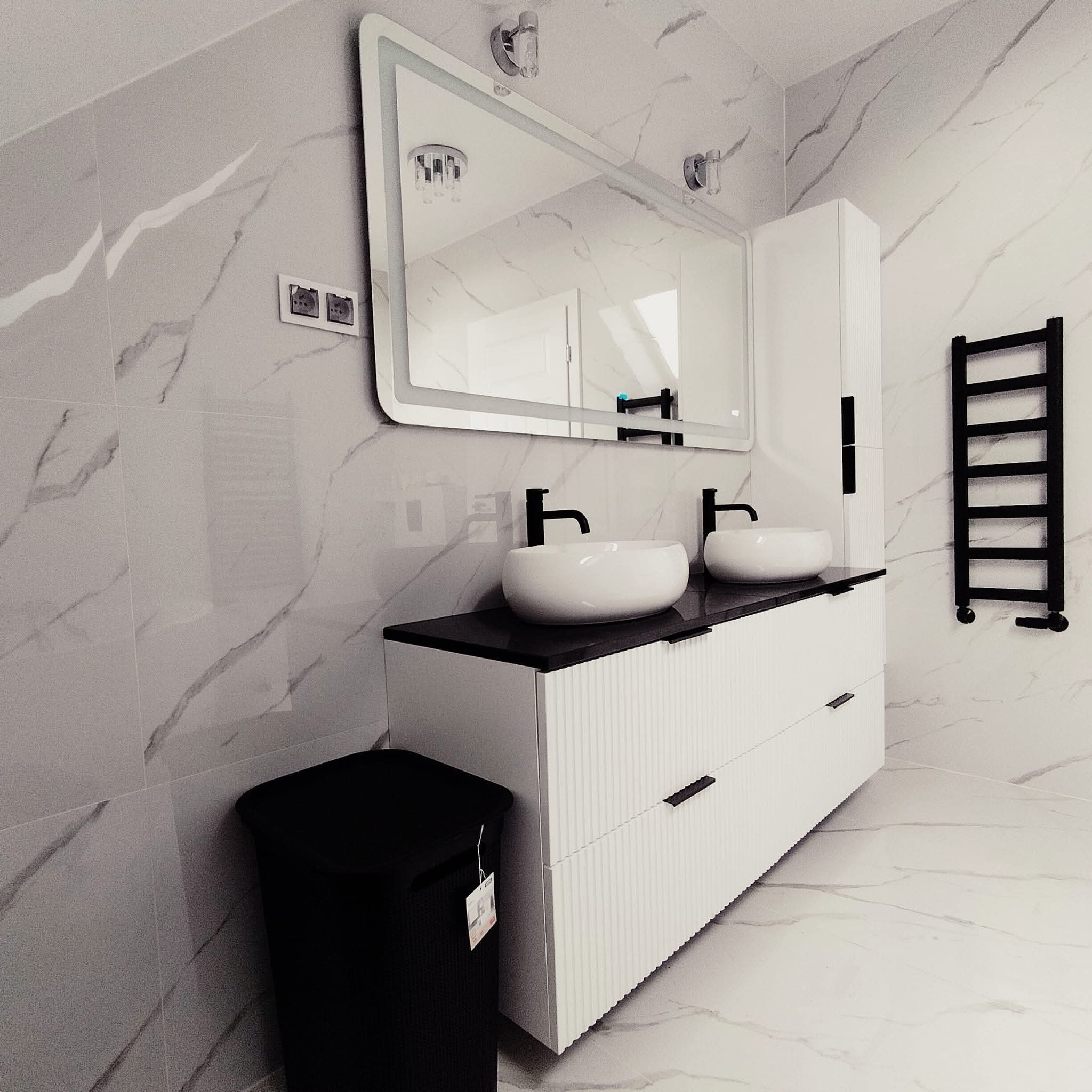 Nowoczesna łazienka z marmurowymi płytkami, białą szafką z umywalkami nablatowymi, podświetlanym lustrem i czarnym grzejnikiem drabinkowym.