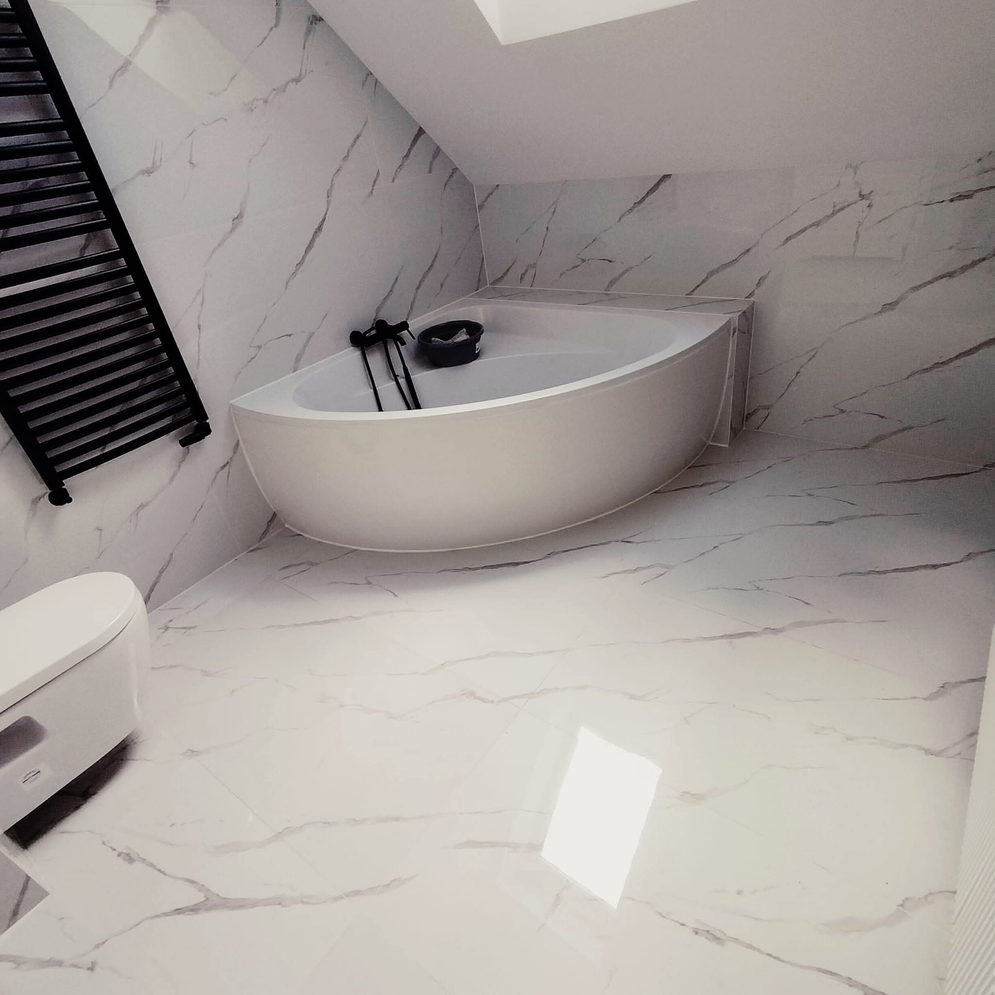 Nowoczesna łazienka w bieli i szarości z marmurowym wzorem na płytkach, owalna wanna, czarny grzejnik drabinkowy i toaleta. Ukośny sufit z oknem dachowym.