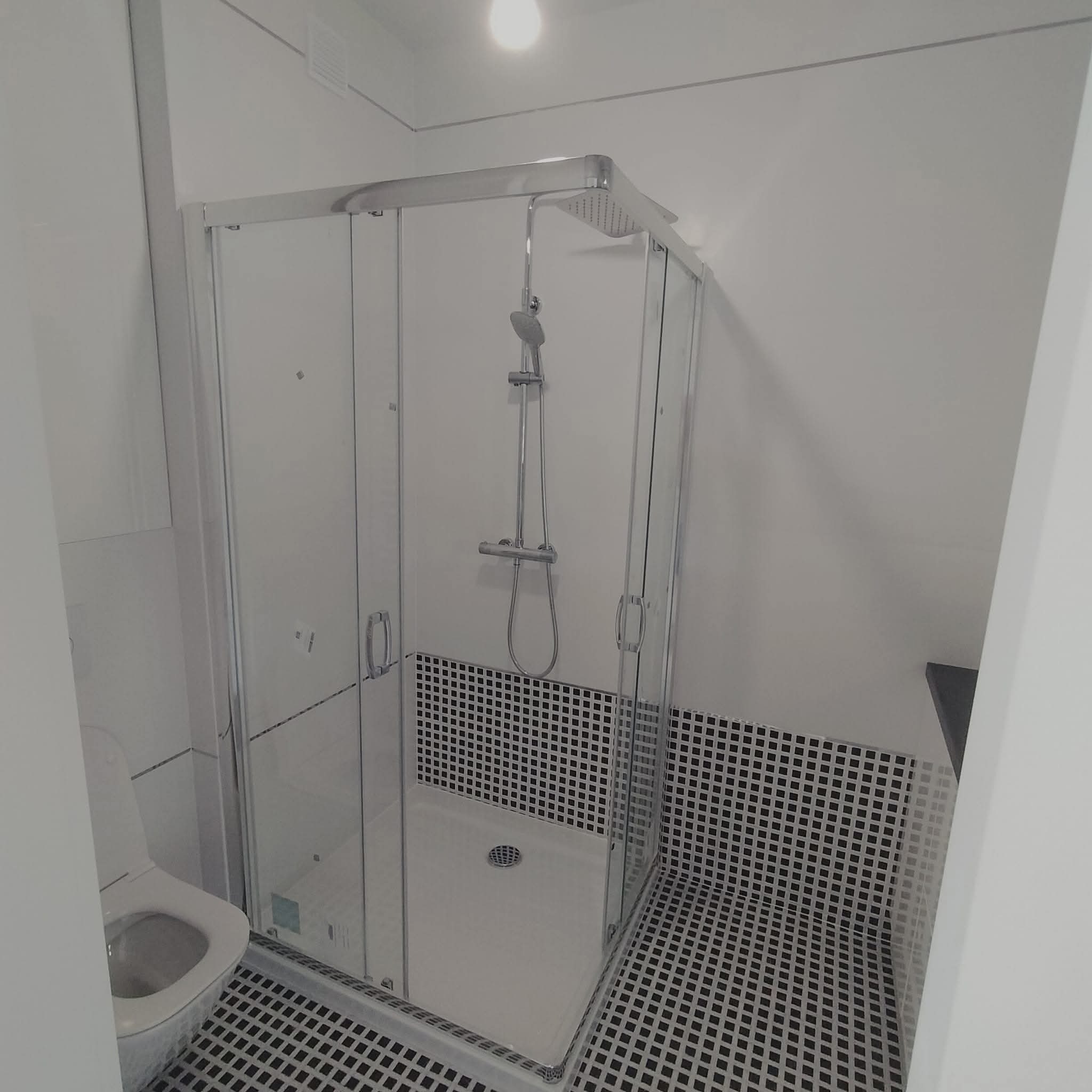 Nowoczesna kabina prysznicowa z czarno-białymi płytkami w łazience. Widoczny fragment toalety. Minimalistyczny design, jasne oświetlenie.