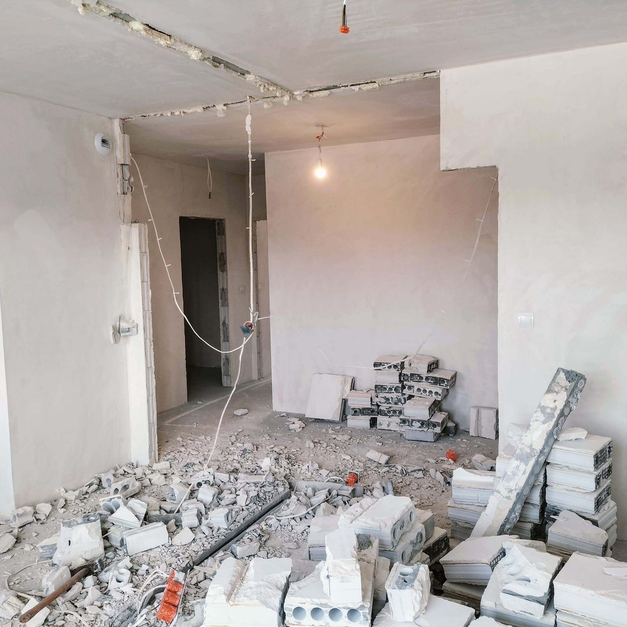 Wnętrze w trakcie remontu: gruz, odsłonięte przewody elektryczne, ściany przygotowane do szpachlowania. Widoczne ślady po demontażu ścianki działowej.