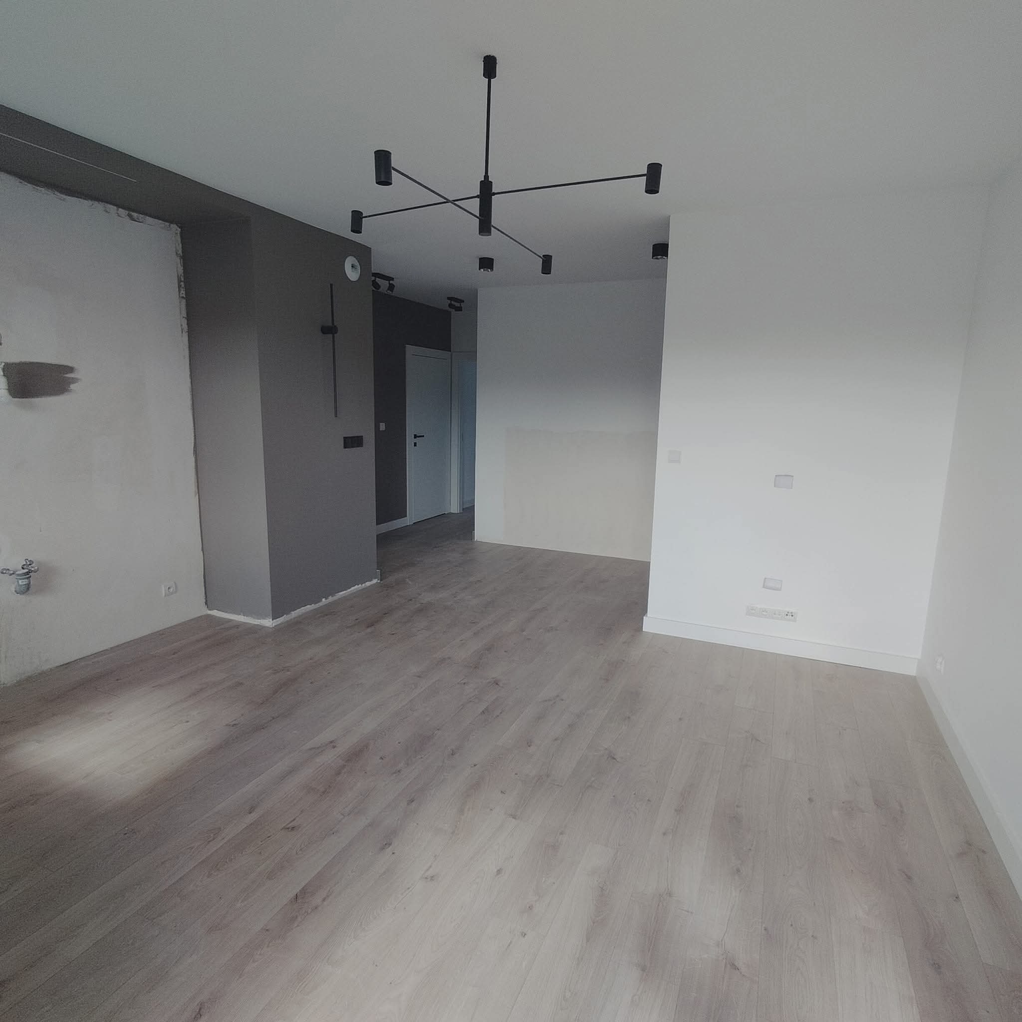 Jasne panele podłogowe w przestronnym, częściowo wykończonym wnętrzu. Ściany w odcieniach bieli i szarości, nowoczesna lampa sufitowa. Widoczne gniazdka elektryczne.