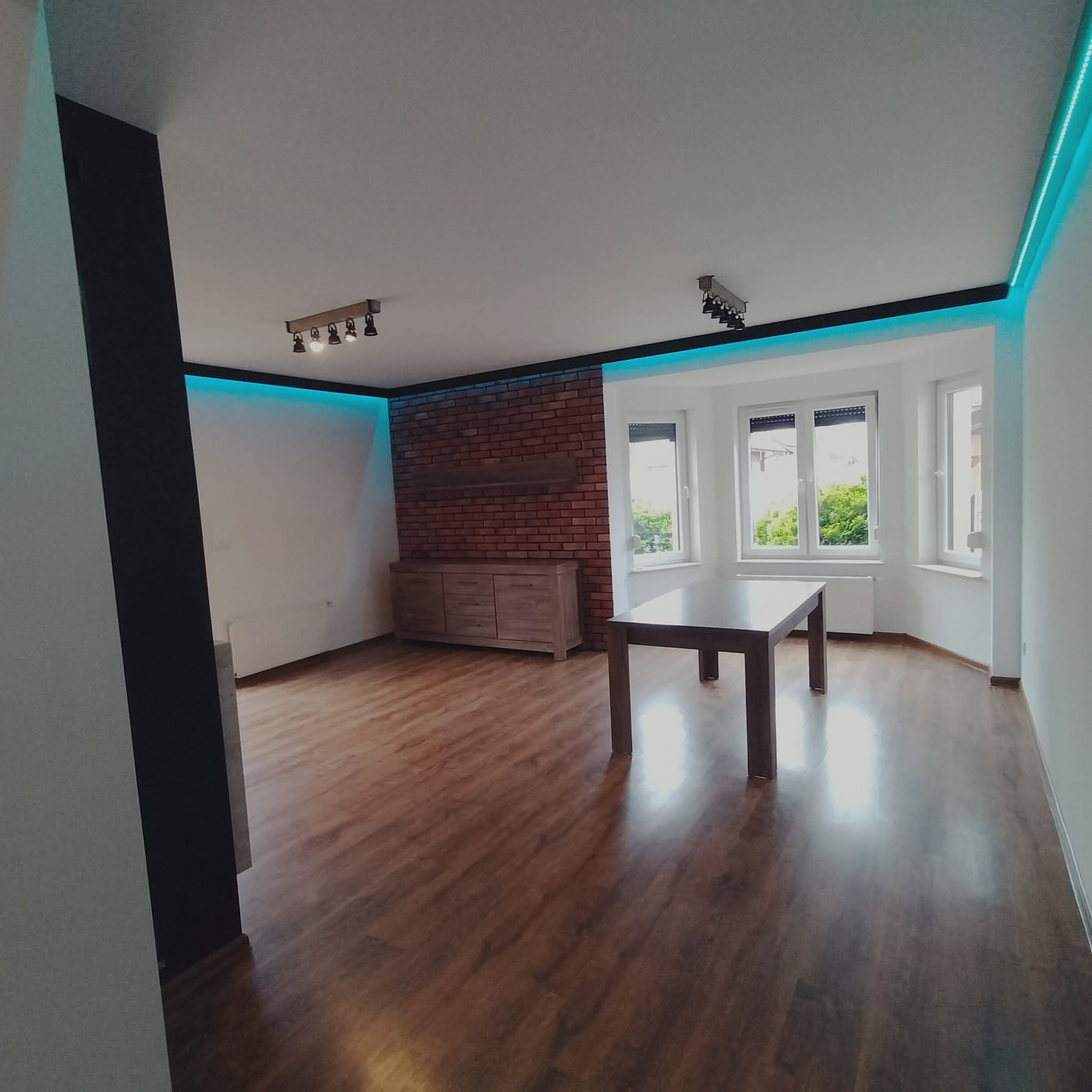Jasne wnętrze po remoncie: drewniana podłoga, ceglana ściana z komodą, stół, okna z widokiem na zieleń, oświetlenie LED. Nowoczesny design z akcentami cegły i drewna.