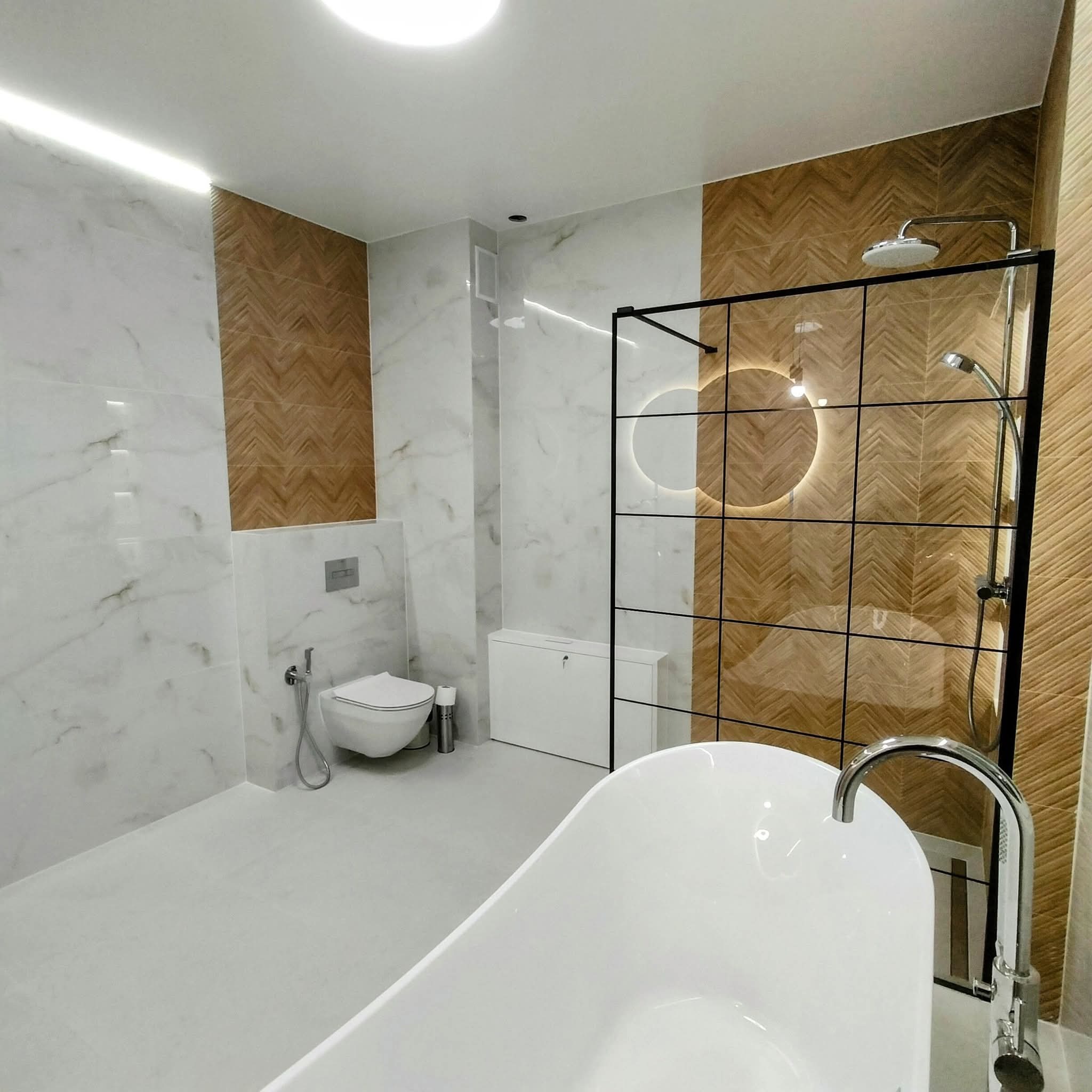 Nowoczesna łazienka z wolnostojącą wanną, kabiną prysznicową typu walk-in z czarnymi profilami i okrągłym lustrem z podświetleniem LED. Biało-brązowe płytki na ścianach.
