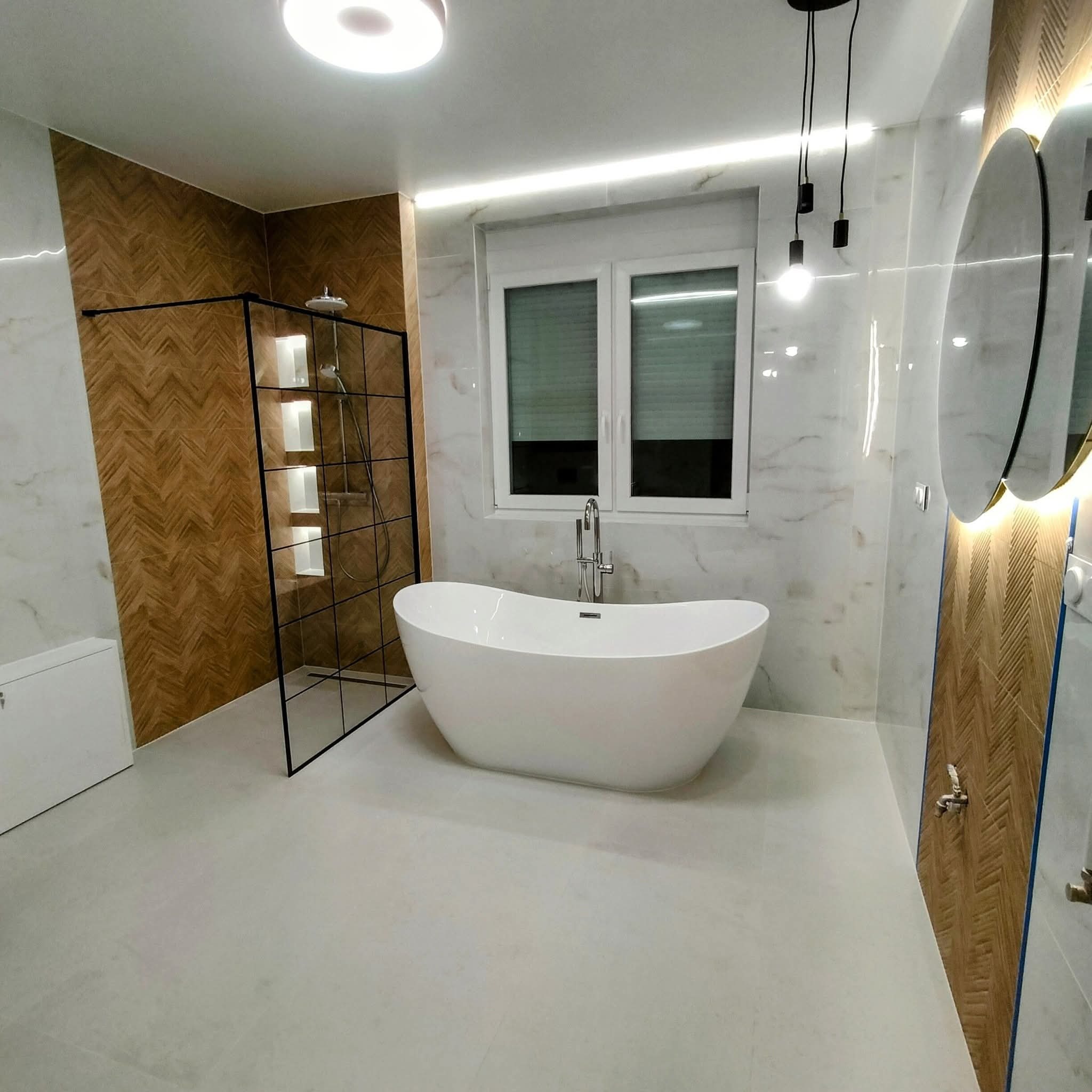 Nowoczesna łazienka z wolnostojącą wanną, kabiną prysznicową typu walk-in z półkami i oświetleniem LED. Marmurowe płytki i drewnopodobna ściana w jodełkę.