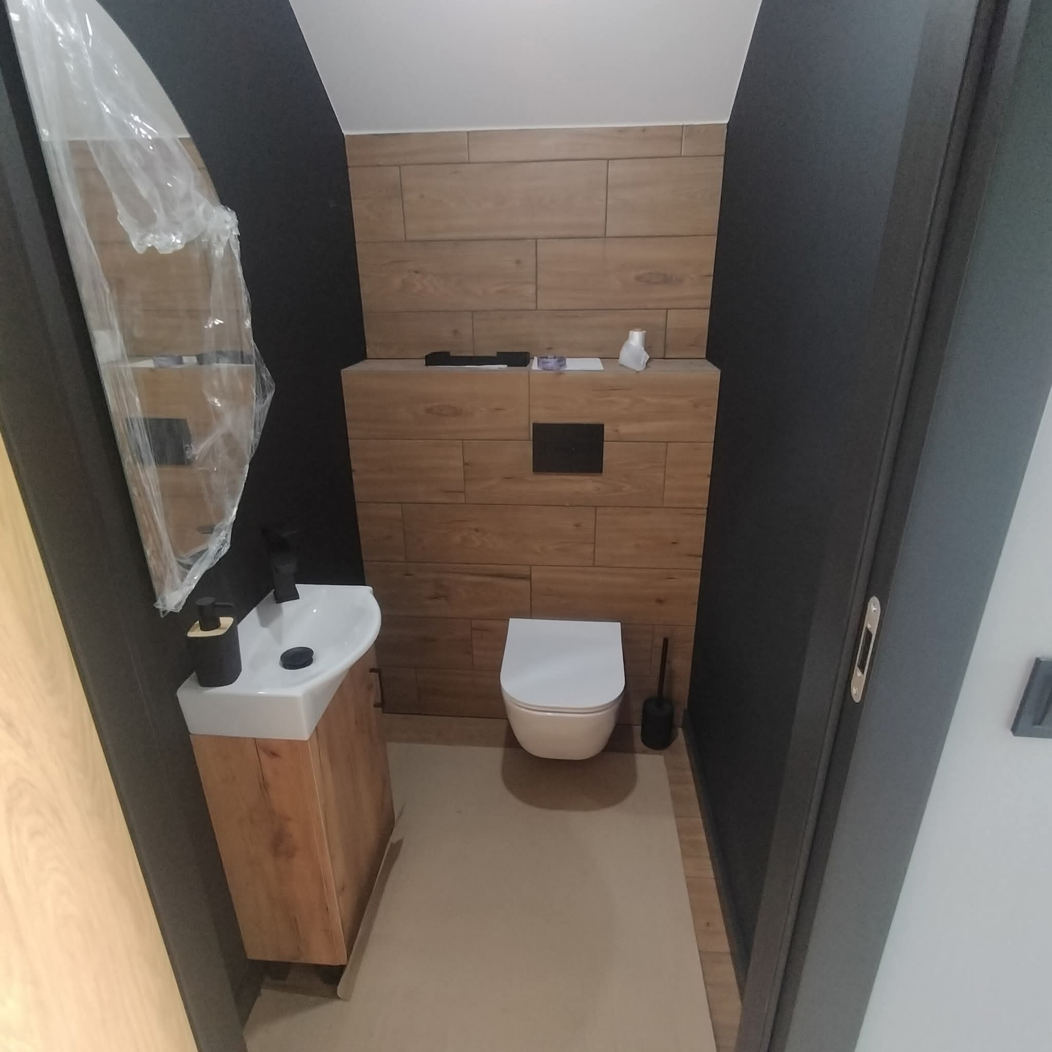 Nowoczesna łazienka po remoncie: drewnopodobne płytki na ścianie, wisząca toaleta, umywalka z szafką w stylu drewna, czarne ściany. Lustro zabezpieczone folią.