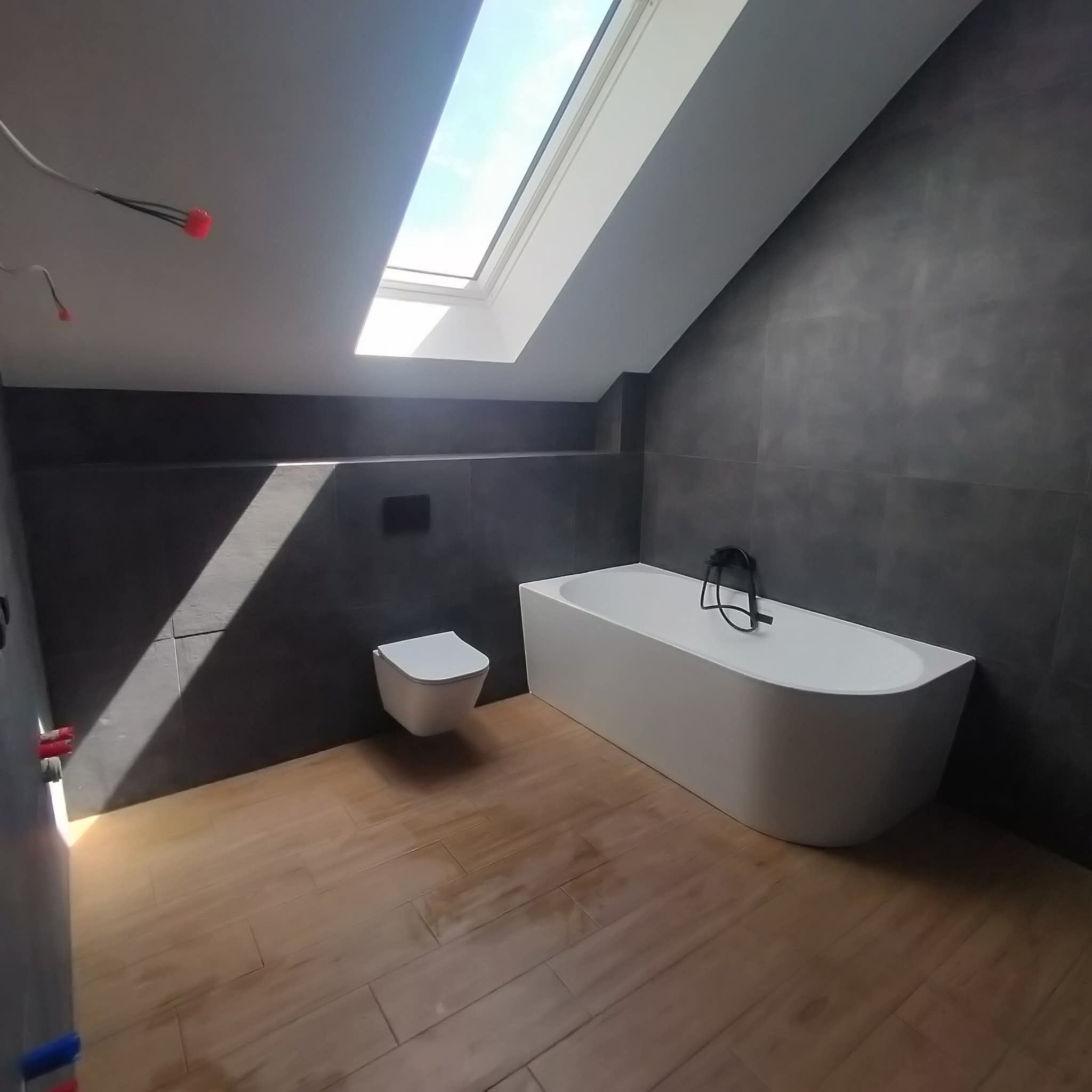 Nowoczesna łazienka na poddaszu z drewnopodobną podłogą, białą wanną i wiszącą toaletą. Okno dachowe wpuszcza naturalne światło. Ciemne płytki na ścianach.