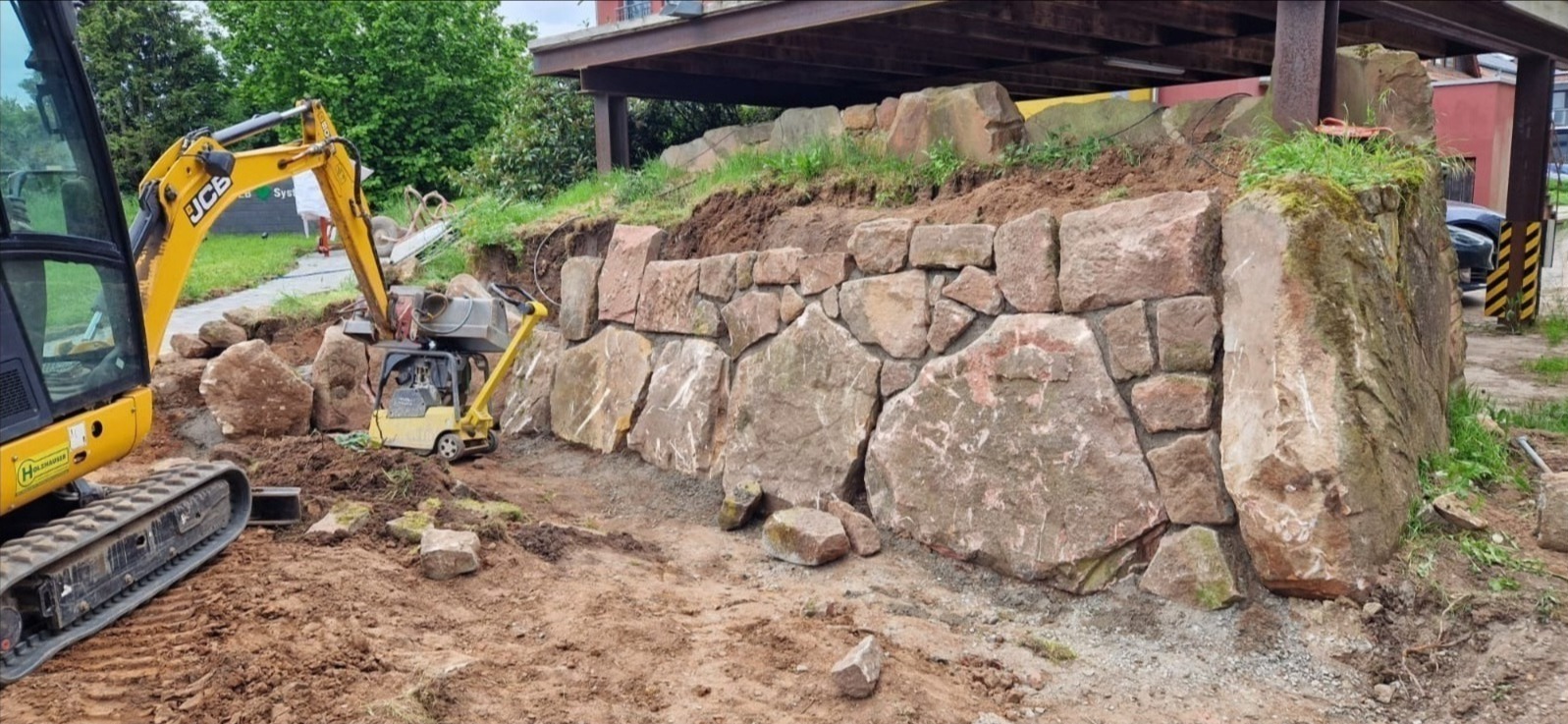 Budowa muru oporowego z kamienia naturalnego. Żółta koparka i zagęszczarka w tle. Prace ziemne w toku, widoczne nierówności terenu i roślinność.