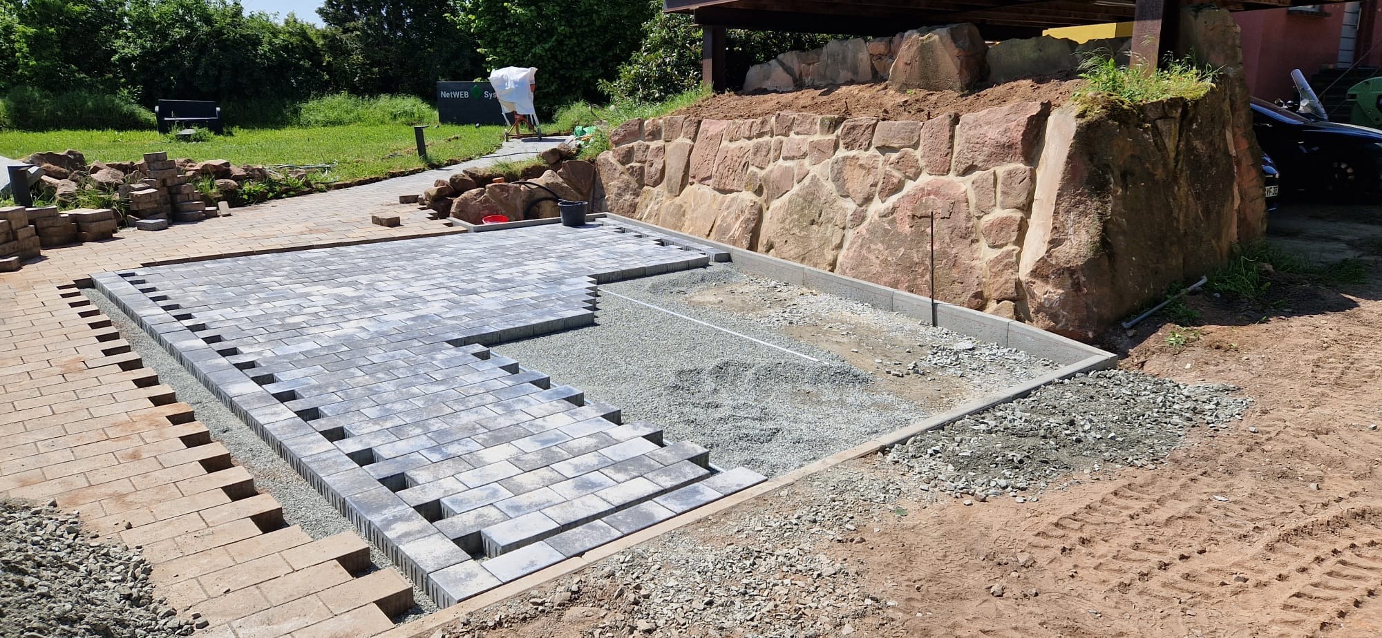 Układanie szarej kostki brukowej na podsypce z grysu, widoczne etapy prac, obrzeża betonowe, w tle mur z kamienia i zieleń. Częściowo ułożona nawierzchnia.