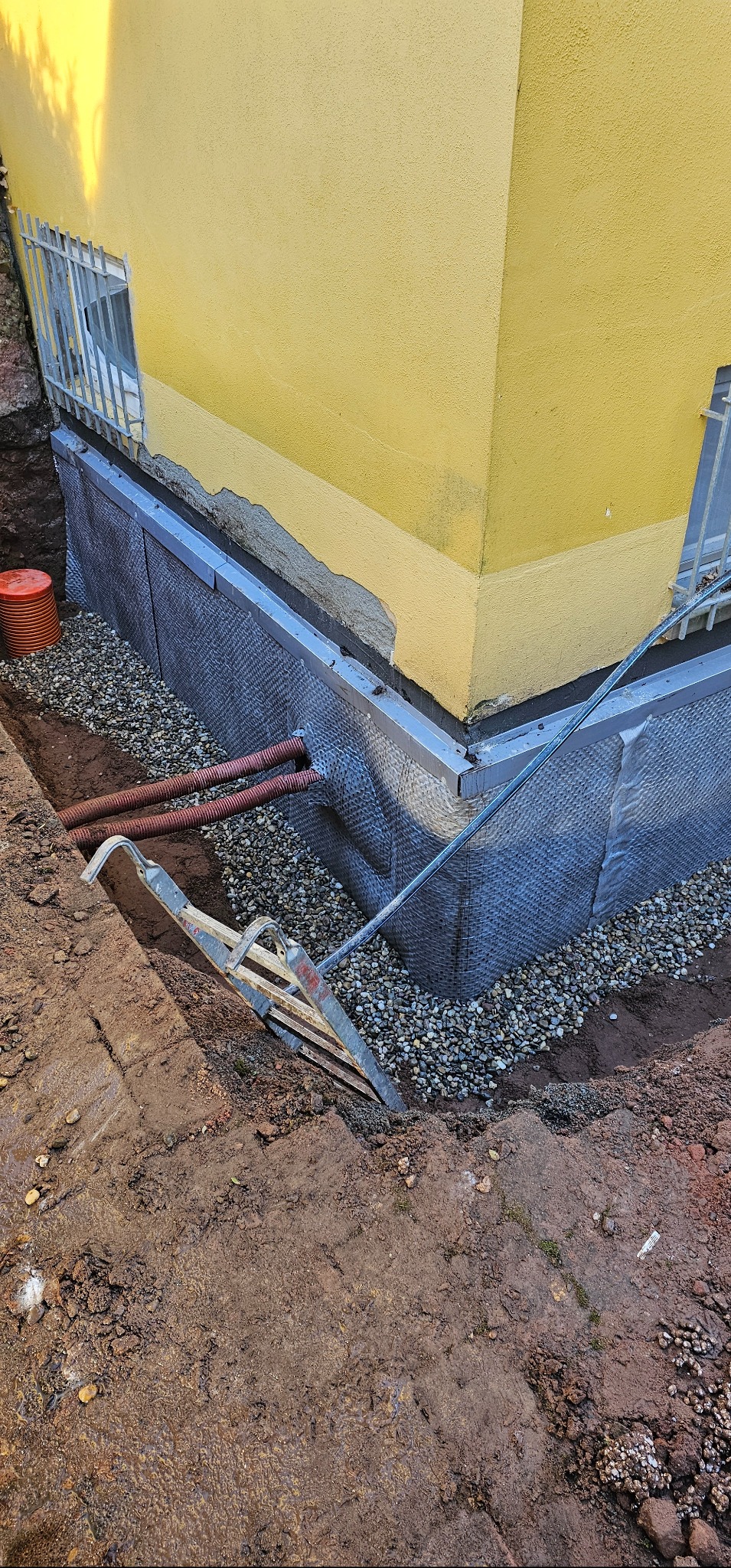 Ocieplenie fundamentu budynku z żółtą elewacją, widoczne rury i warstwa izolacyjna z siatką. W tle okno z kratą i ziemia.