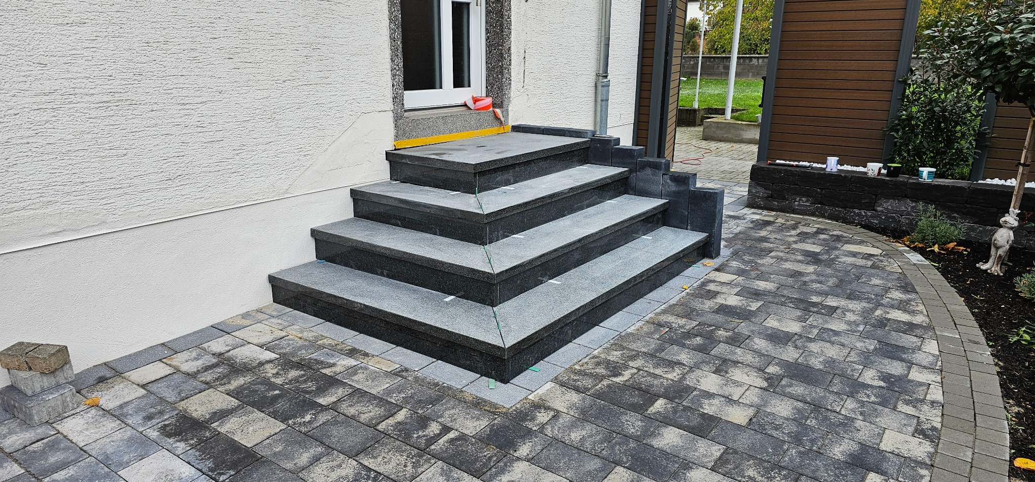 Granitowe schody wejściowe z kostką brukową w odcieniach szarości, prowadzące do drzwi z oknem. Widoczne elementy wykończeniowe i dekoracyjne.