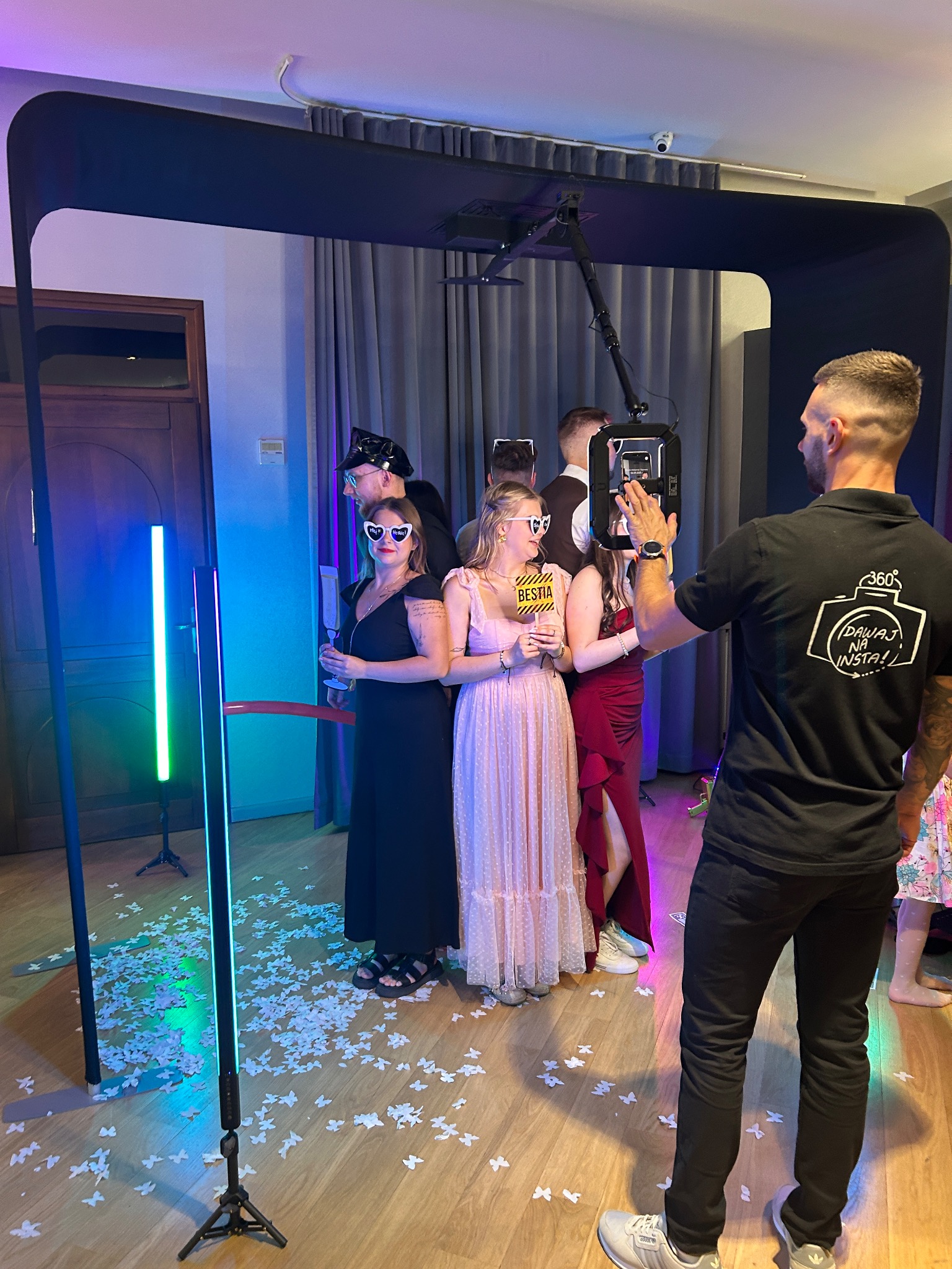 Uczestnicy imprezy pozują do zdjęcia w fotobudce 360 z rekwizytami, konfetti na podłodze, obsługa w tle. Dynamiczne ujęcie z imprezy okolicznościowej.