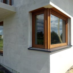 Docieplenia Elewacje Wojciech - Szara elewacja domu jednorodzinnego z drewnianą stolarką okienną i czarnym parapetem, widok z zewnątrz, fragment budynku z oknem narożnym.