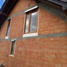 Docieplenia Elewacje Wojciech - Surowa elewacja z czerwonej cegły ceramicznej z zamontowanymi oknami z brązowymi ramami. Widoczne betonowe nadproża i fragment dachu. Budynek w trakcie budowy.