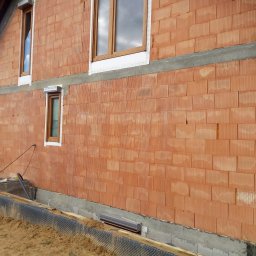 Docieplenia Elewacje Wojciech - Ściana domu w trakcie budowy z czerwonej cegły, widoczne okna z drewnianymi ramami i fundamenty. Etap murowania ścian.