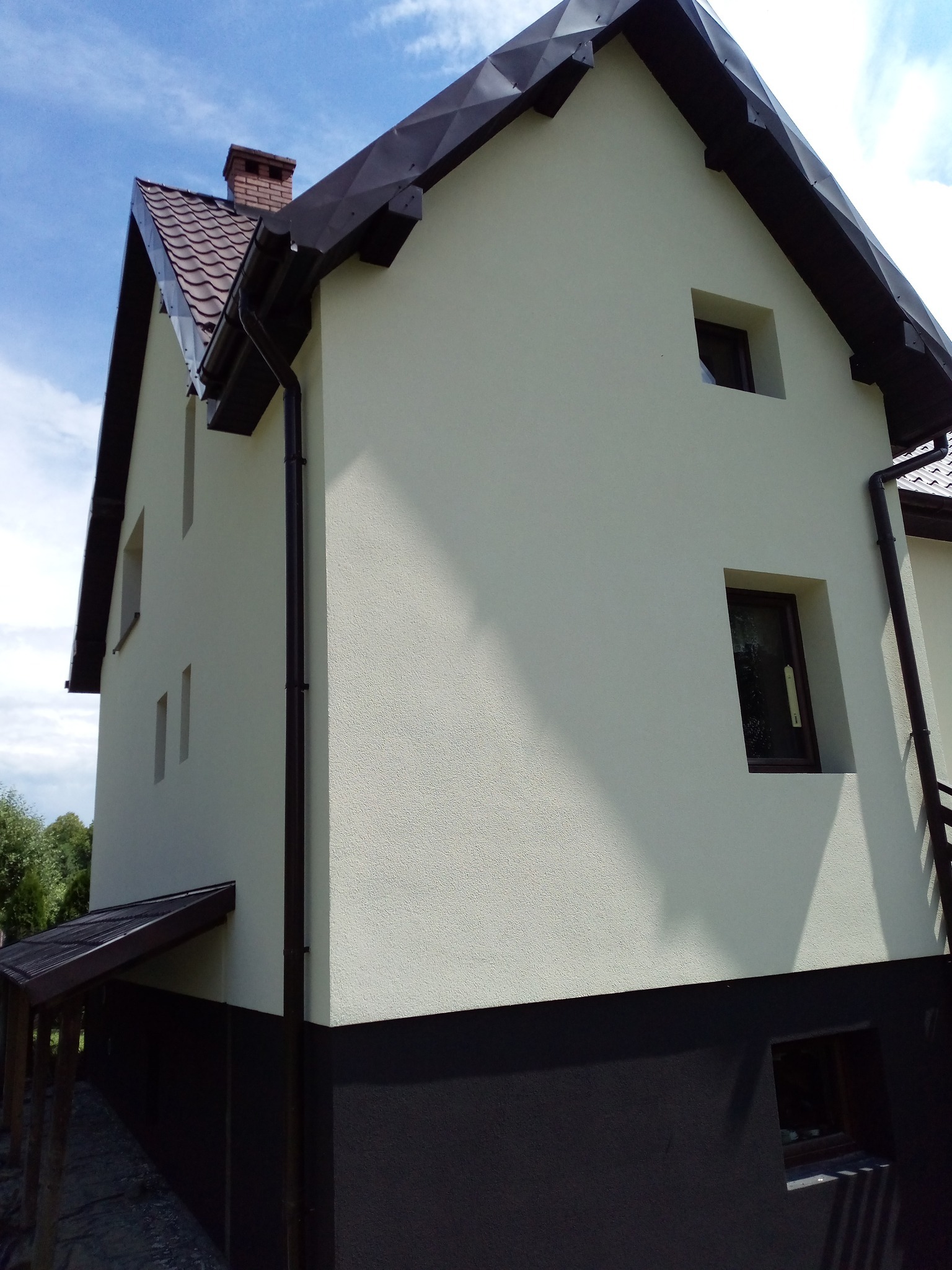 Dwukondygnacyjny dom z jasną elewacją i ciemnym cokołem, widoczny z boku pod kątem. Ciemna stolarka okienna i dach z brązowymi rynnami. Błękitne niebo w tle.