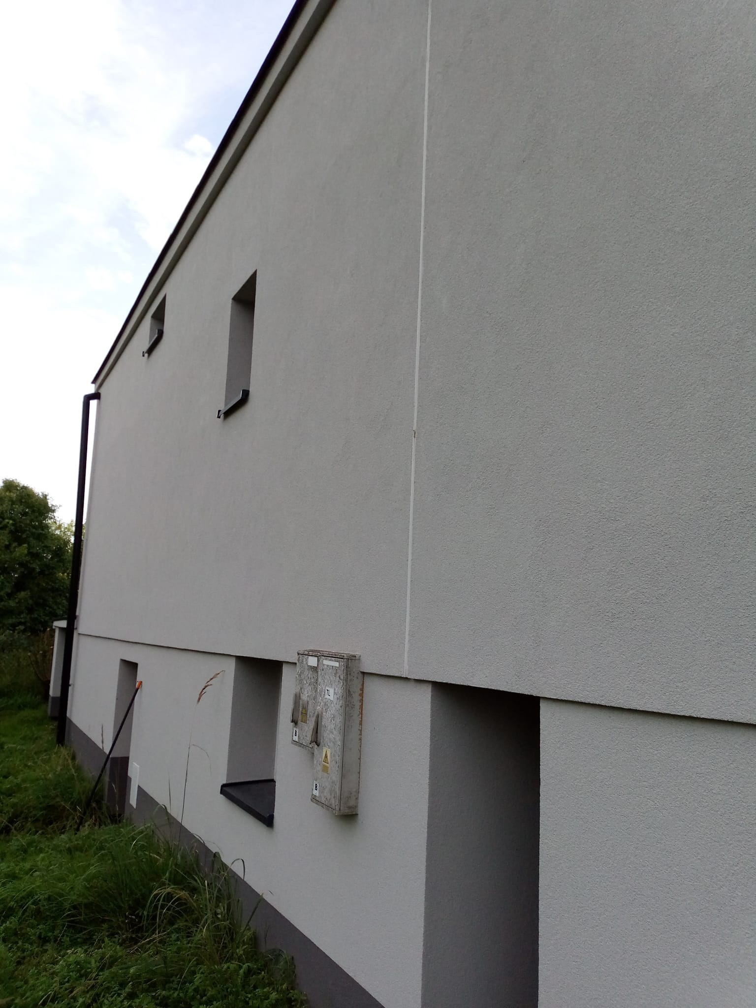 Jasnoszara elewacja budynku z widocznymi skrzynkami elektrycznymi i małymi oknami. Świeżo otynkowana ściana z widoczną fakturą tynku.