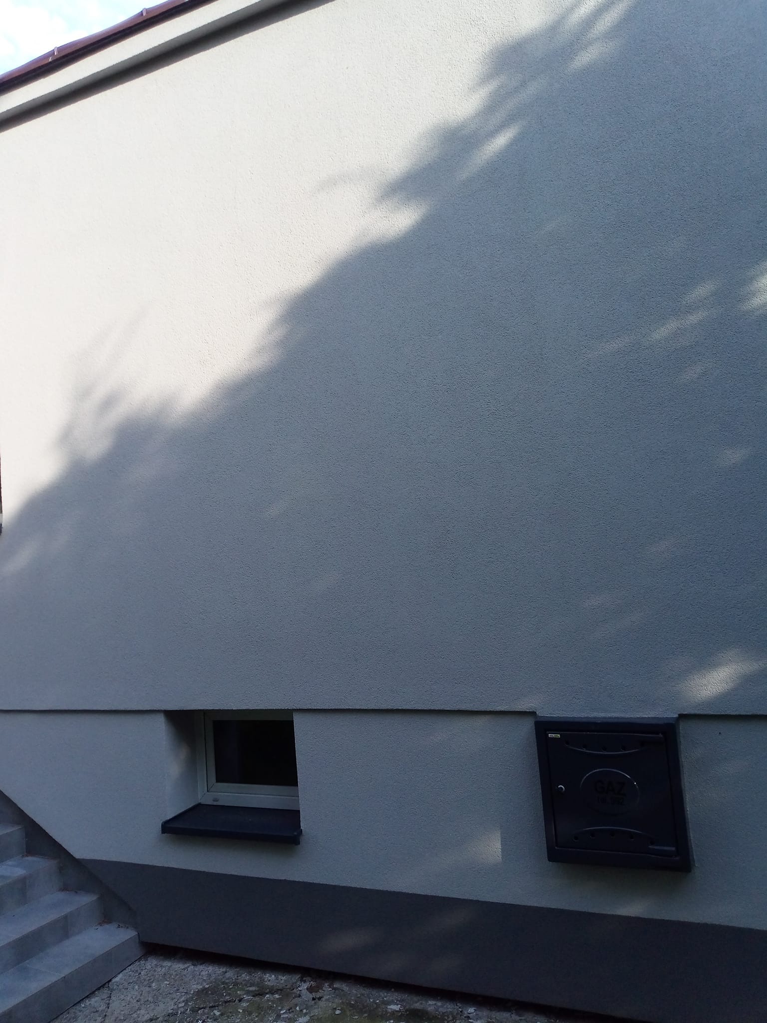 Jasnoszara elewacja budynku z widocznym oknem piwnicznym, skrzynką gazową i fragmentem schodów. Na ścianie cień drzewa. Tekstura tynku barankowego.