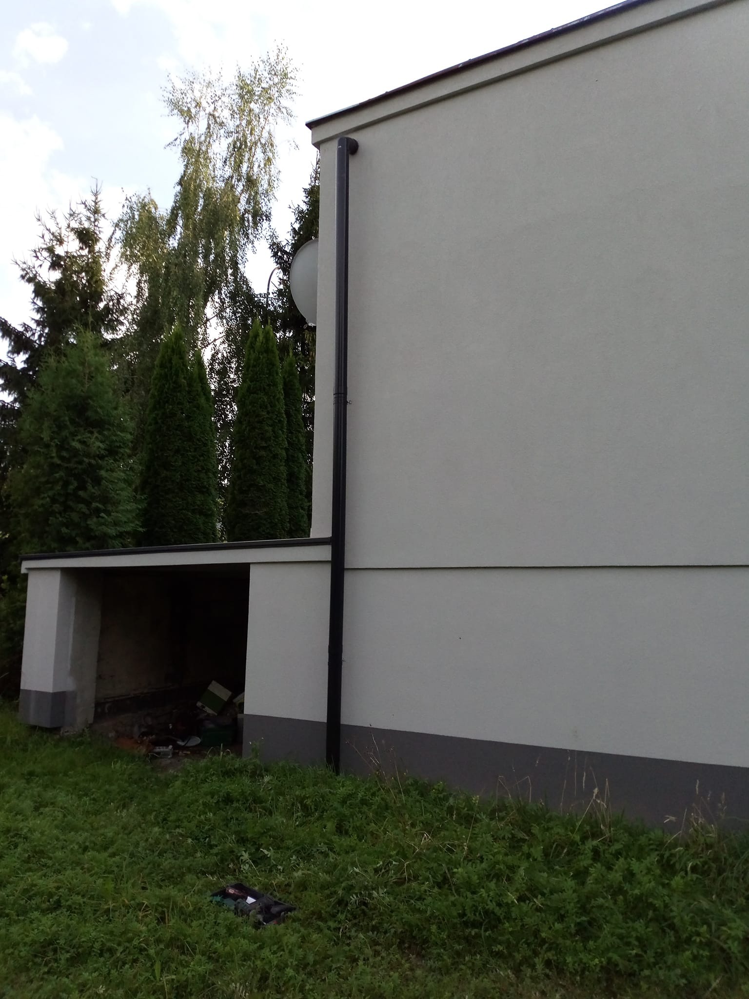 Jasna elewacja budynku z garażem, widoczna rynna i antena satelitarna. W tle drzewa iglaste i liściaste. Ujęcie z poziomu trawy.