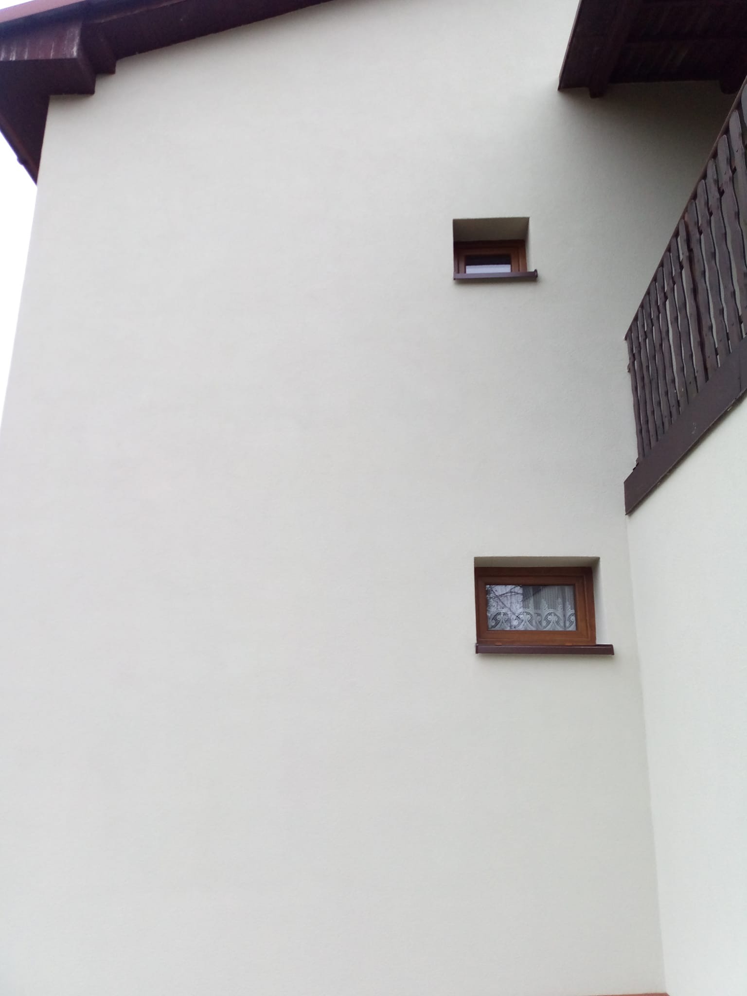 Jasna elewacja budynku z dwoma oknami o brązowych ramach i fragmentem drewnianej balustrady balkonu. Minimalistyczny design, neutralna kolorystyka.