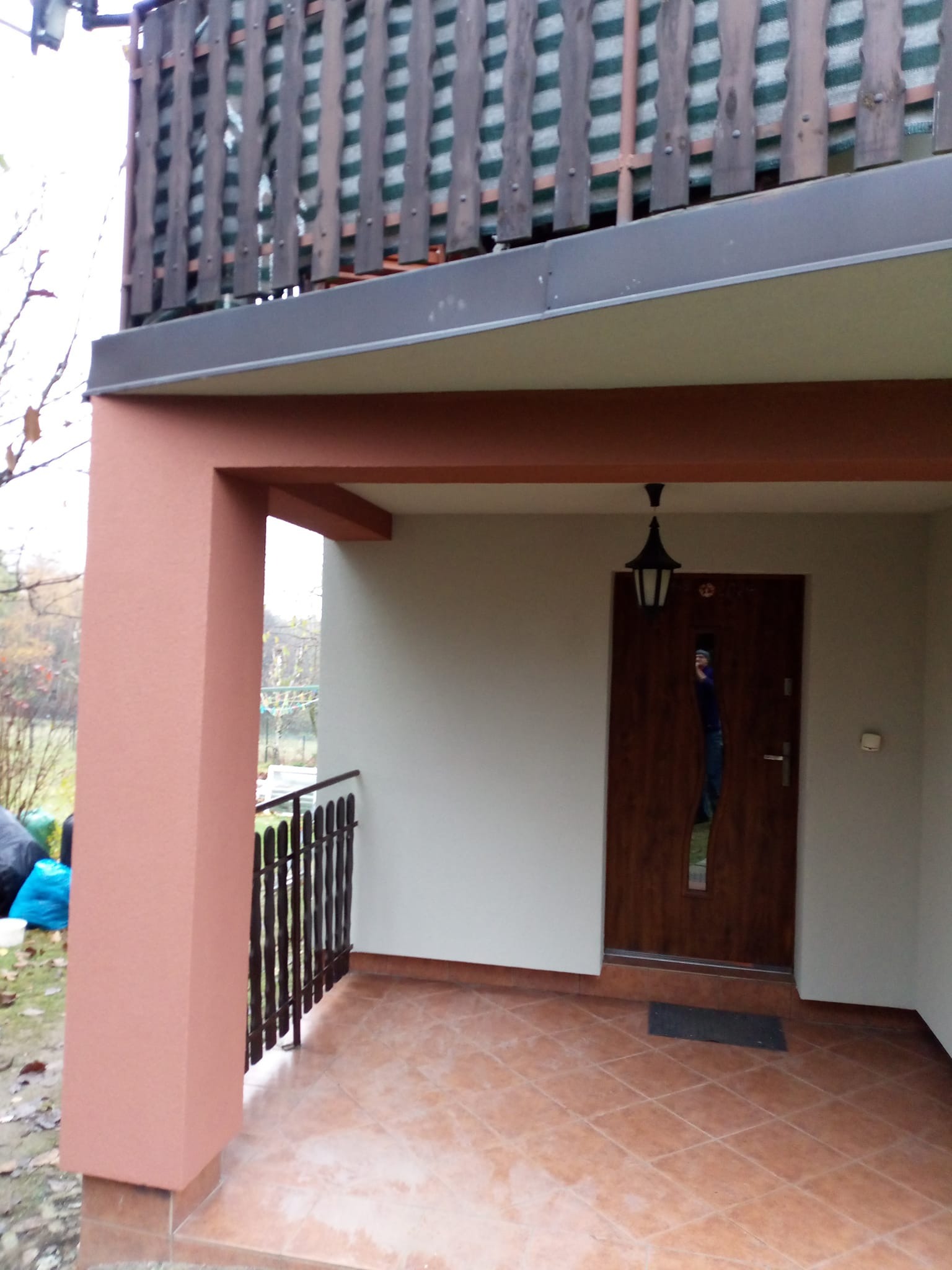 Wykończony, zadaszony taras z brązową kolumną i balustradą. Ściany w jasnym odcieniu, brązowe płytki na podłodze. Na górze drewniana balustrada balkonu.