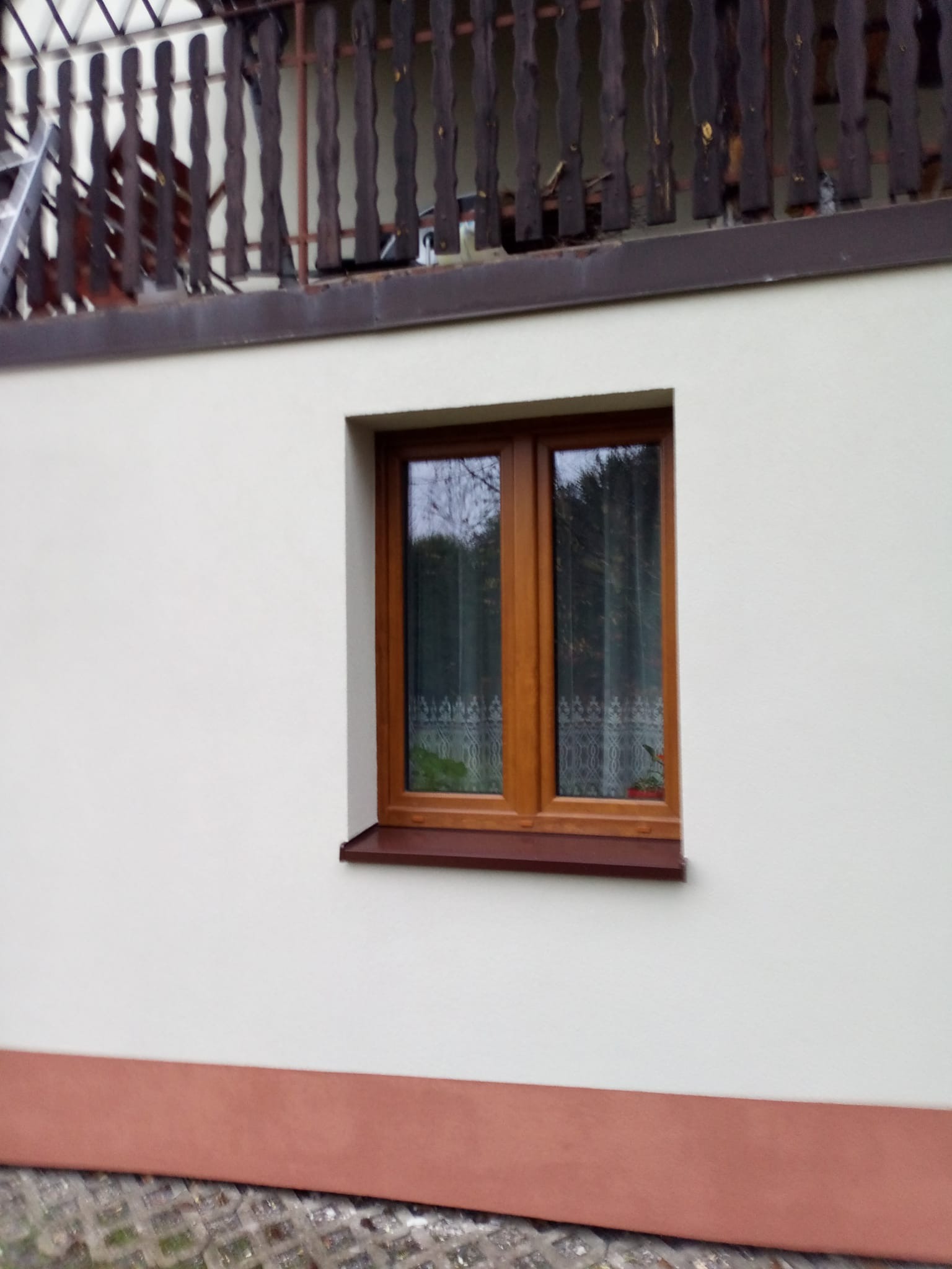 Otynkowana, kremowa elewacja domu z brązowym oknem i cokołem. Widoczny fragment balkonu z drewnianą balustradą. Ujęcie z dołu, podkreślające detale wykończenia.