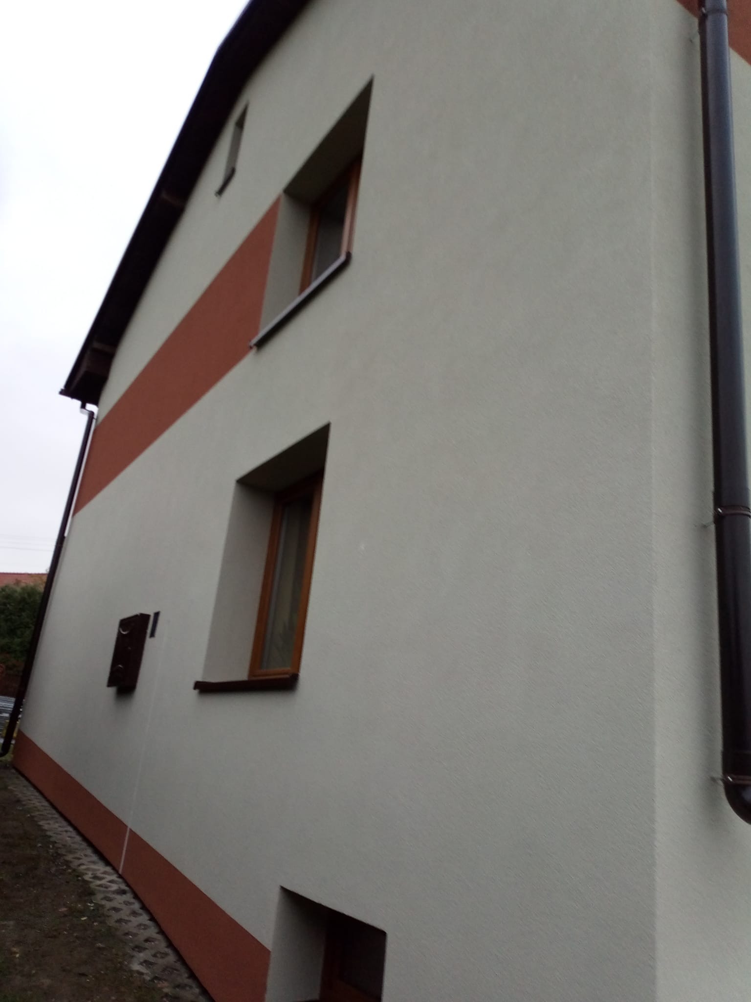 Ściana budynku po tynkowaniu i malowaniu z brązowymi akcentami, widoczne okna z drewnianymi ramami i rynna. Ujęcie z dołu, pochmurne niebo w tle.