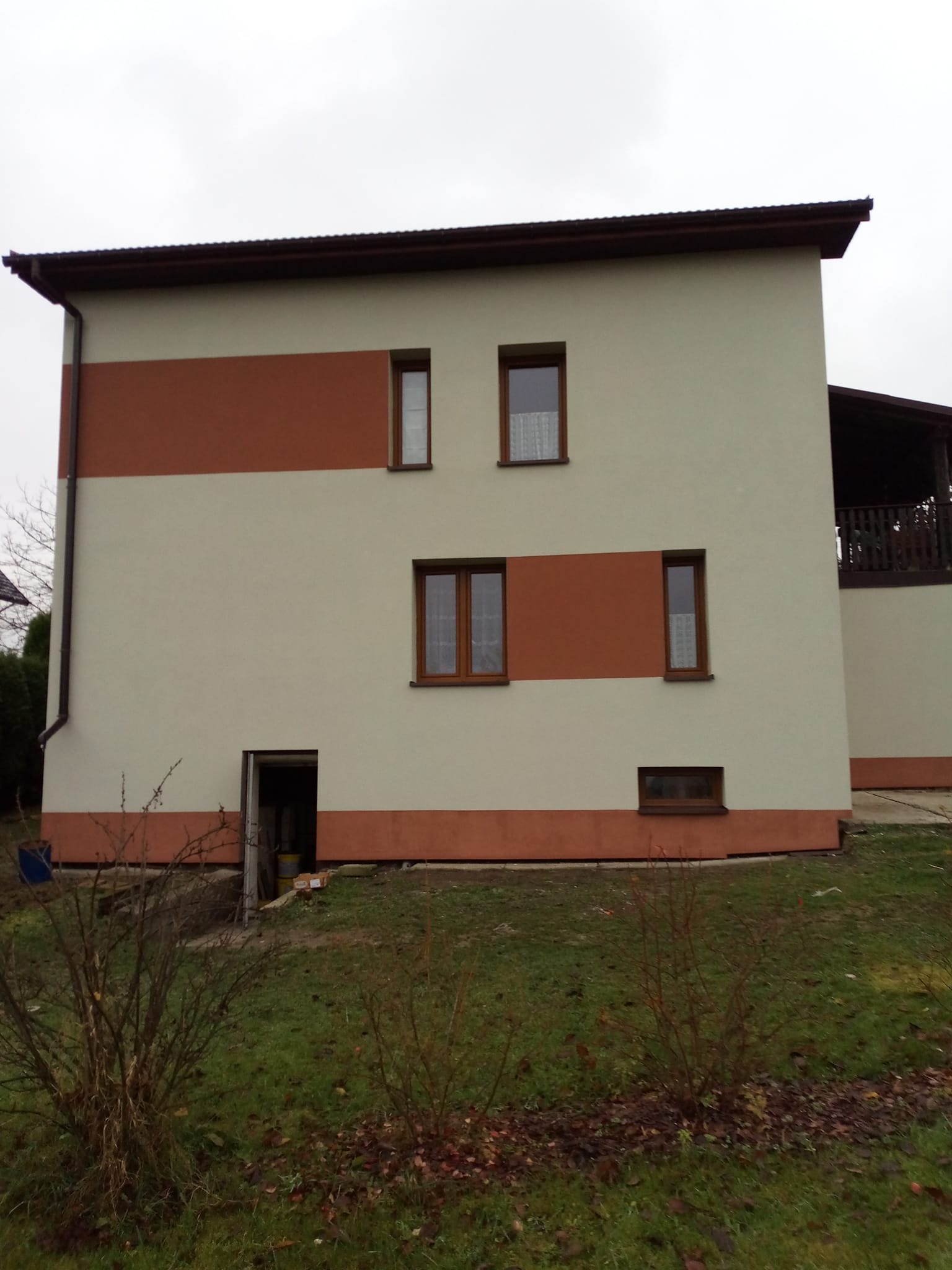 Dwukondygnacyjny dom z elewacją w odcieniach beżu i brązu, z widocznymi oknami z drewnianą stolarką i wejściem do piwnicy. Ujęcie z poziomu gruntu.