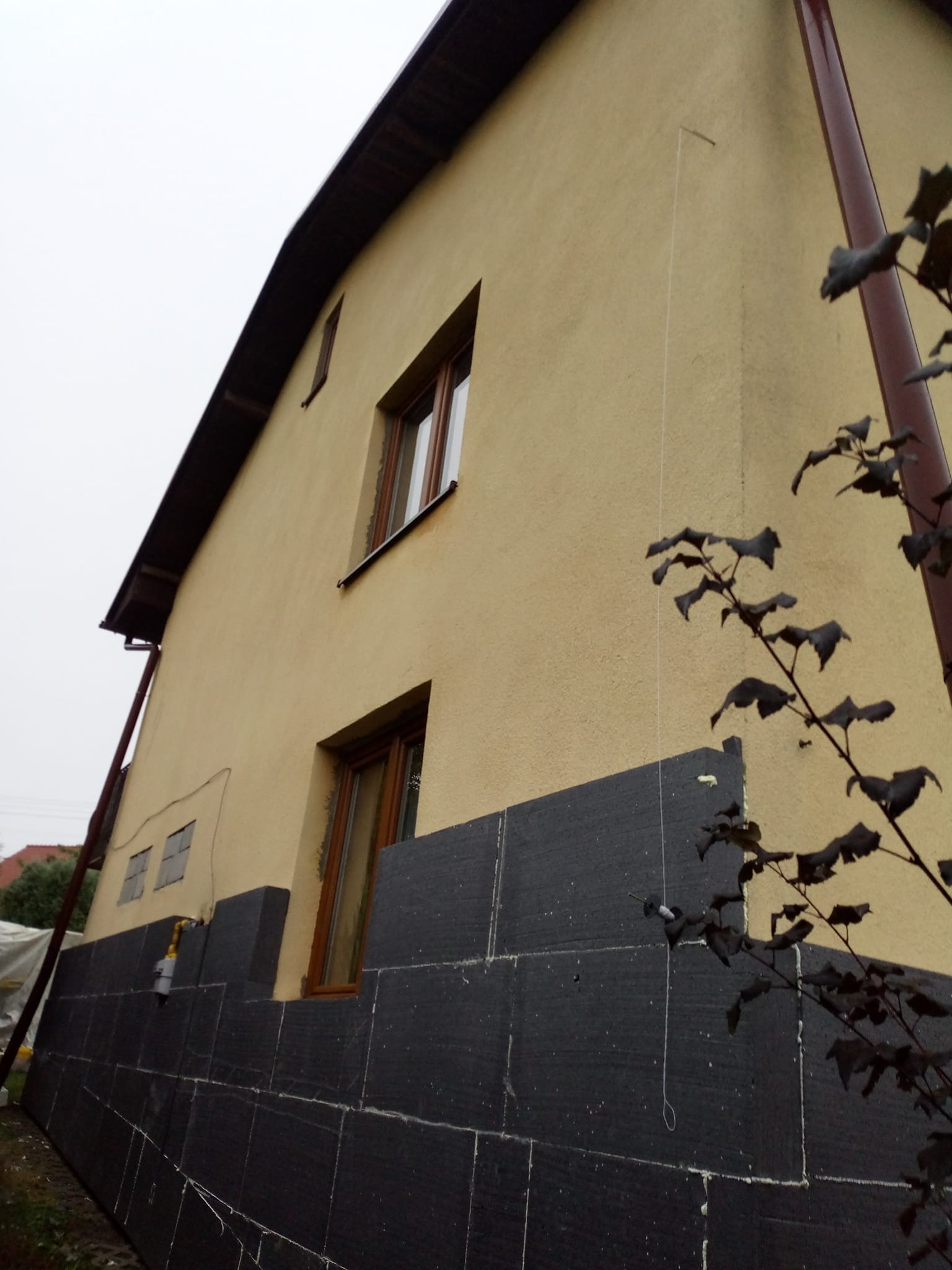Dom w trakcie prac: dolna część ściany pokryta czarnym styropianem, górna w kolorze beżowym. Widoczne okna i drabina oparta o ścianę. Ujęcie z dołu, pochmurne niebo.