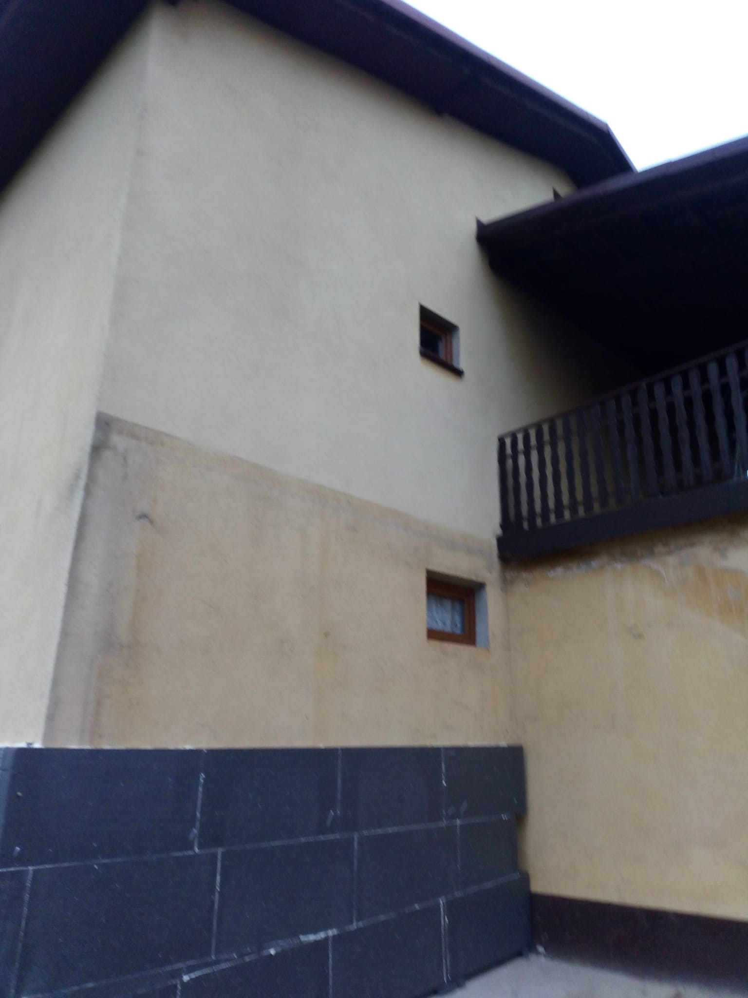 Fragment elewacji budynku z widocznym ociepleniem w dolnej części. Górna część elewacji w odcieniach beżu, z dwoma oknami i balkonem z drewnianą balustradą.