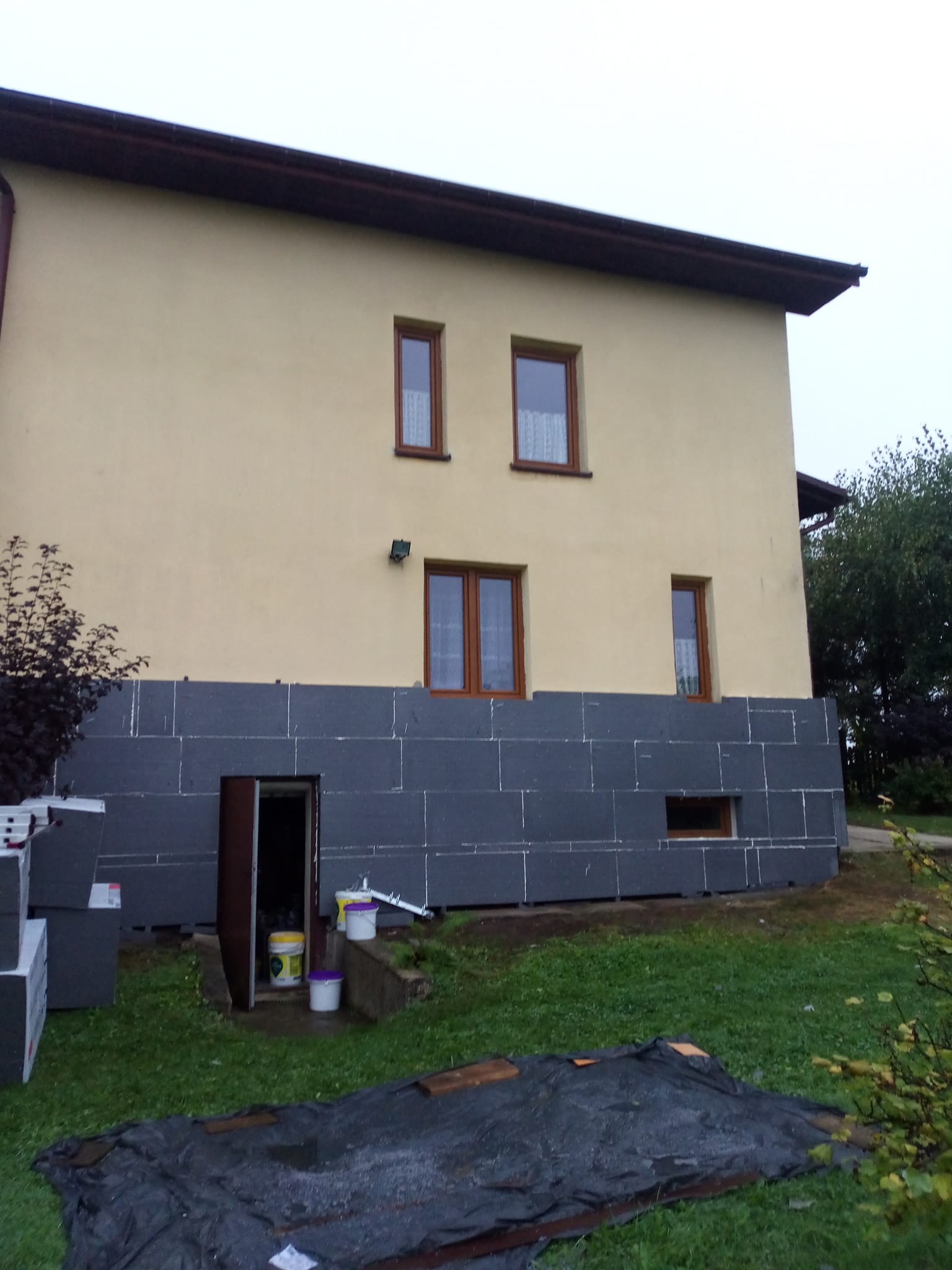 Dom z częściowo ocieploną elewacją styropianem grafitowym. Widoczne puszki z farbą i narzędzia. Prace w toku, dolna część budynku w szarym styropianie.
