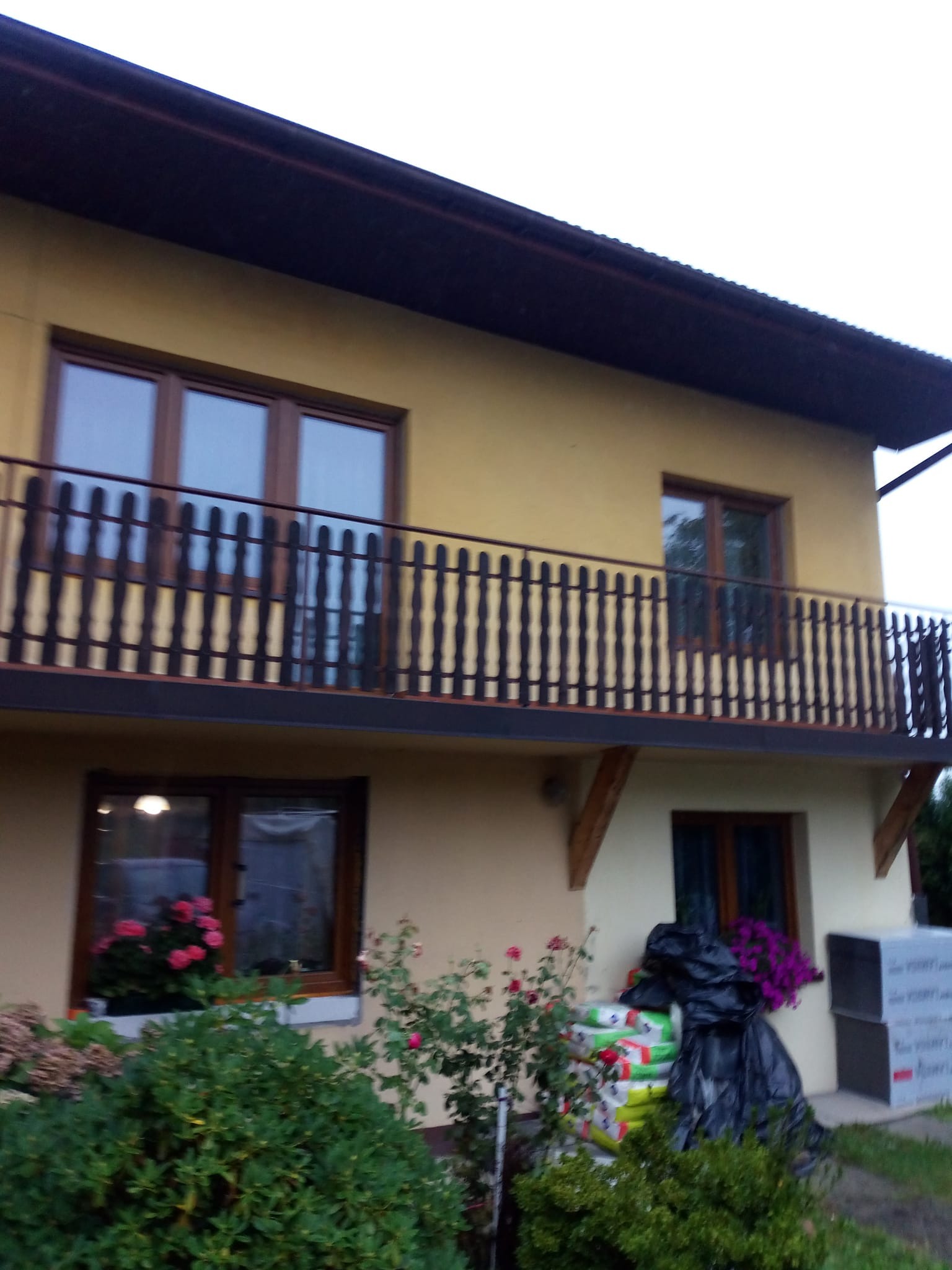 Dwukondygnacyjny dom z balkonem i żółtą elewacją. Widoczne okna z drewnianymi ramami, rośliny w donicach i worki z materiałami budowlanymi przy ścianie.