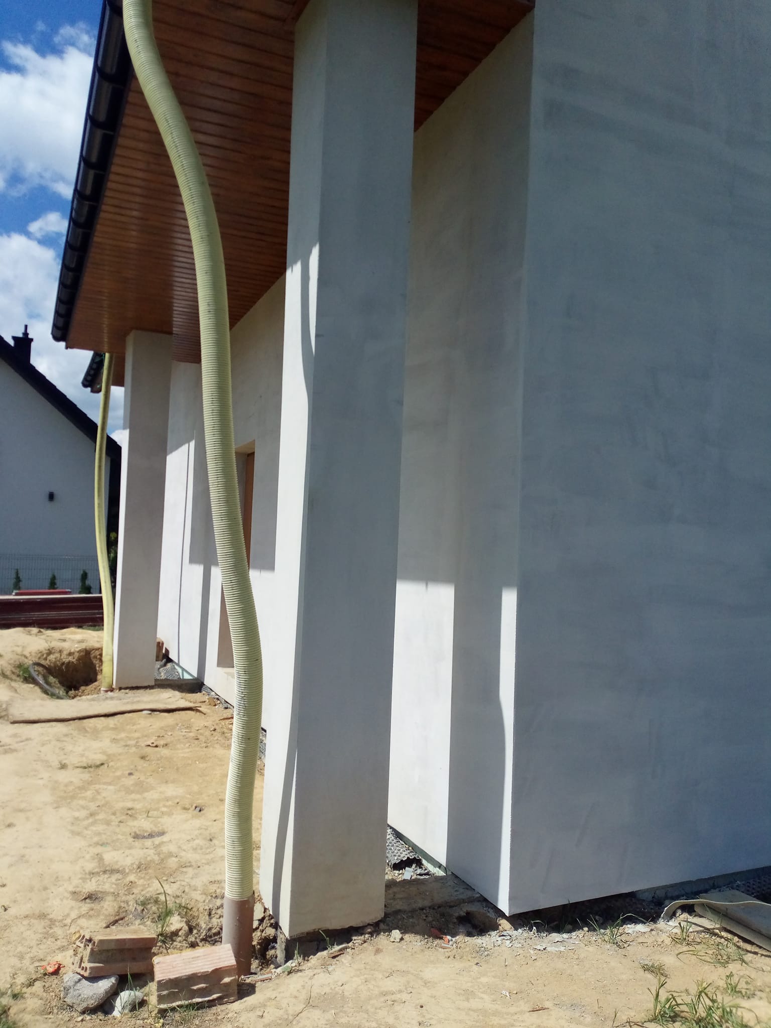 Surowa elewacja domu jednorodzinnego z widocznymi kolumnami i rurą wentylacyjną, w trakcie prac wykończeniowych na zewnątrz budynku. Jasne tynki.