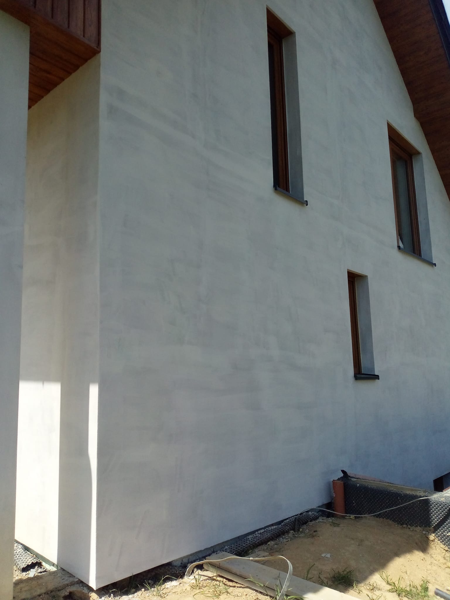 Jasnoszara elewacja domu jednorodzinnego z drewnianymi oknami, widoczna z boku pod kątem. Świeżo położony tynk z delikatną fakturą. Widoczne detale wykończenia.